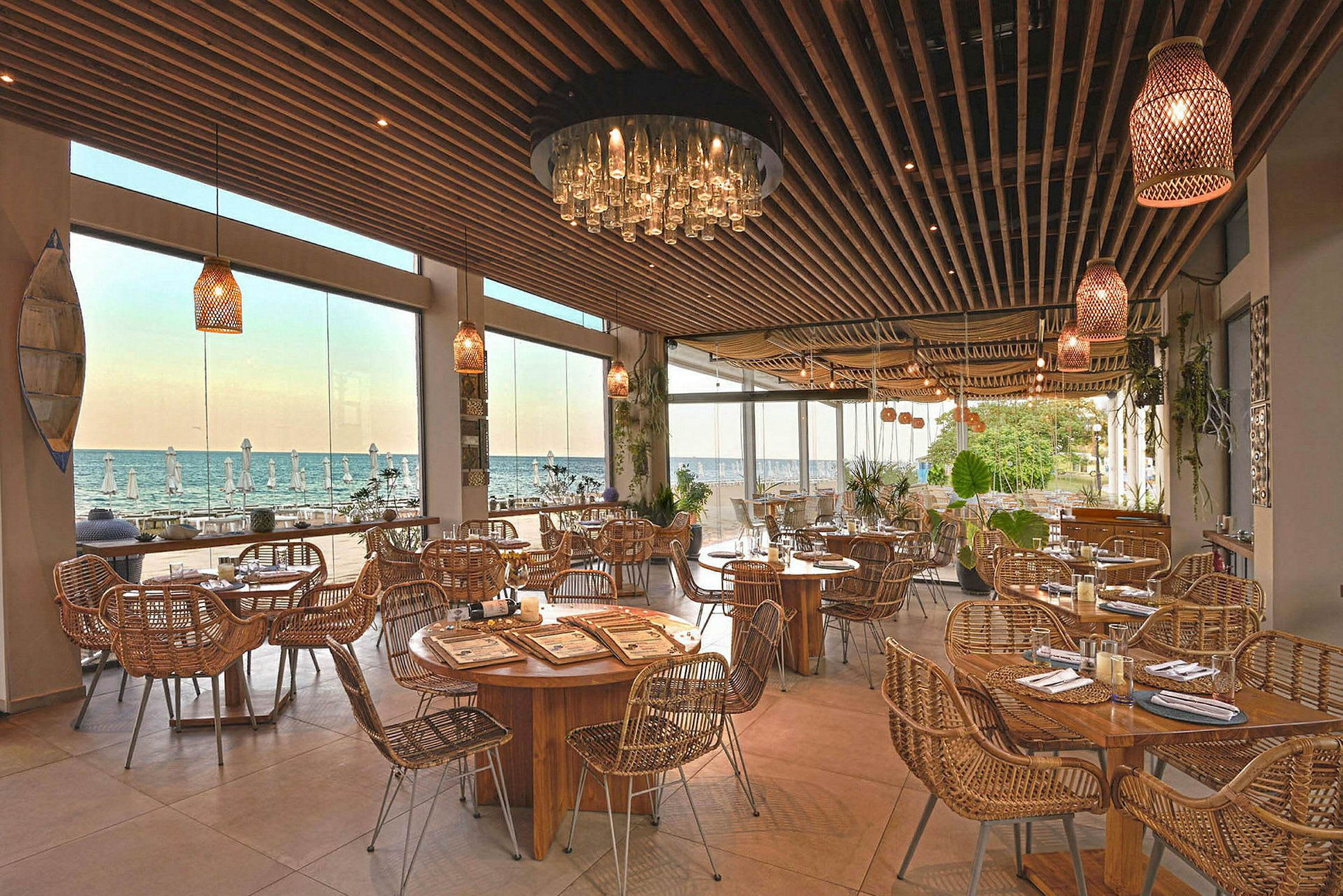 Restaurant med træborde og rottingstole, store vinduer viser udsigt til havet. (AI-genereret tekst)