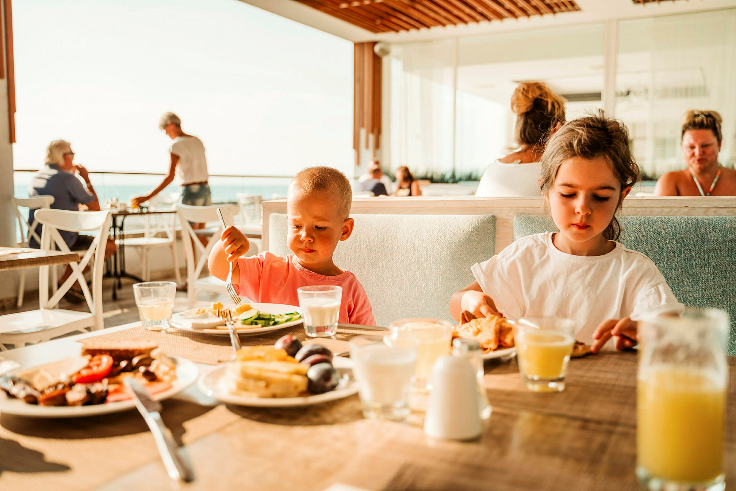 Børn sidder ved et bord og spiser, mens mennesker ses i baggrunden på en restaurant med udsigt. (AI-genereret tekst)