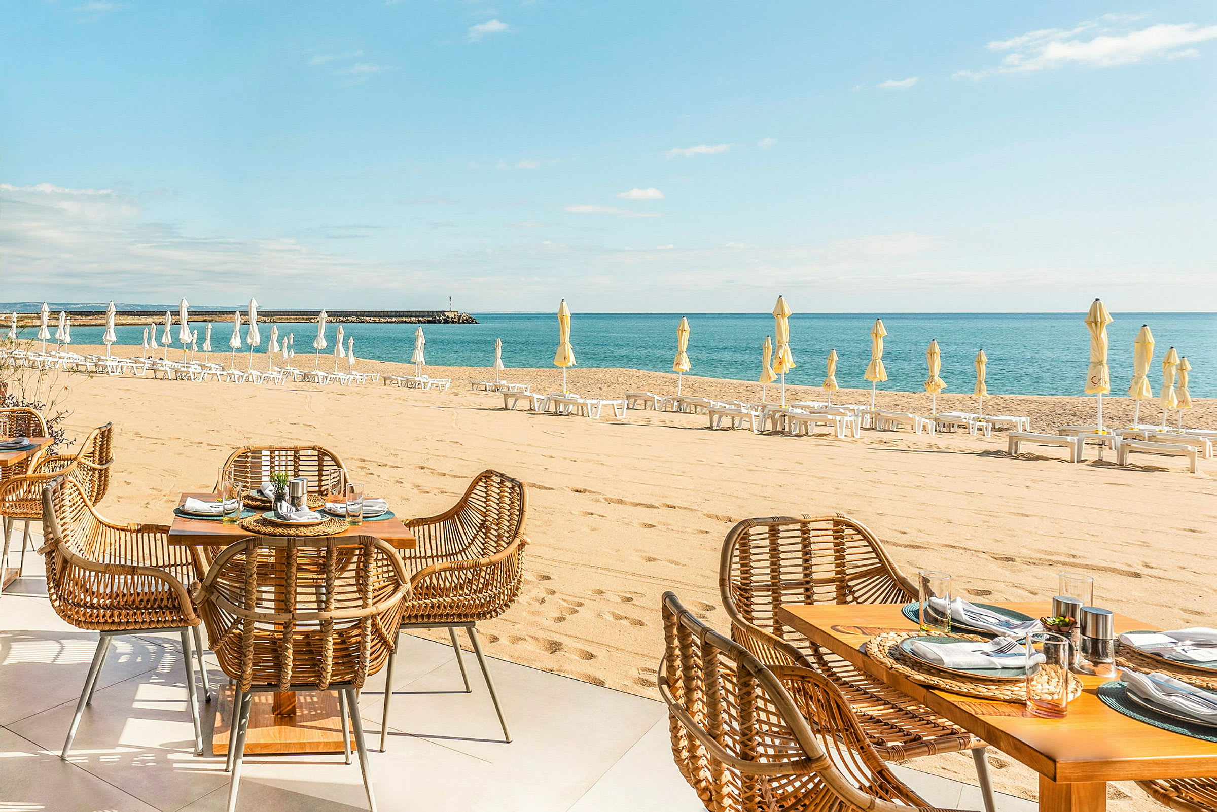 En strand med liggestole og parasoller samt en restaurant med udendørsmøbler i rattan. (AI-genereret tekst)