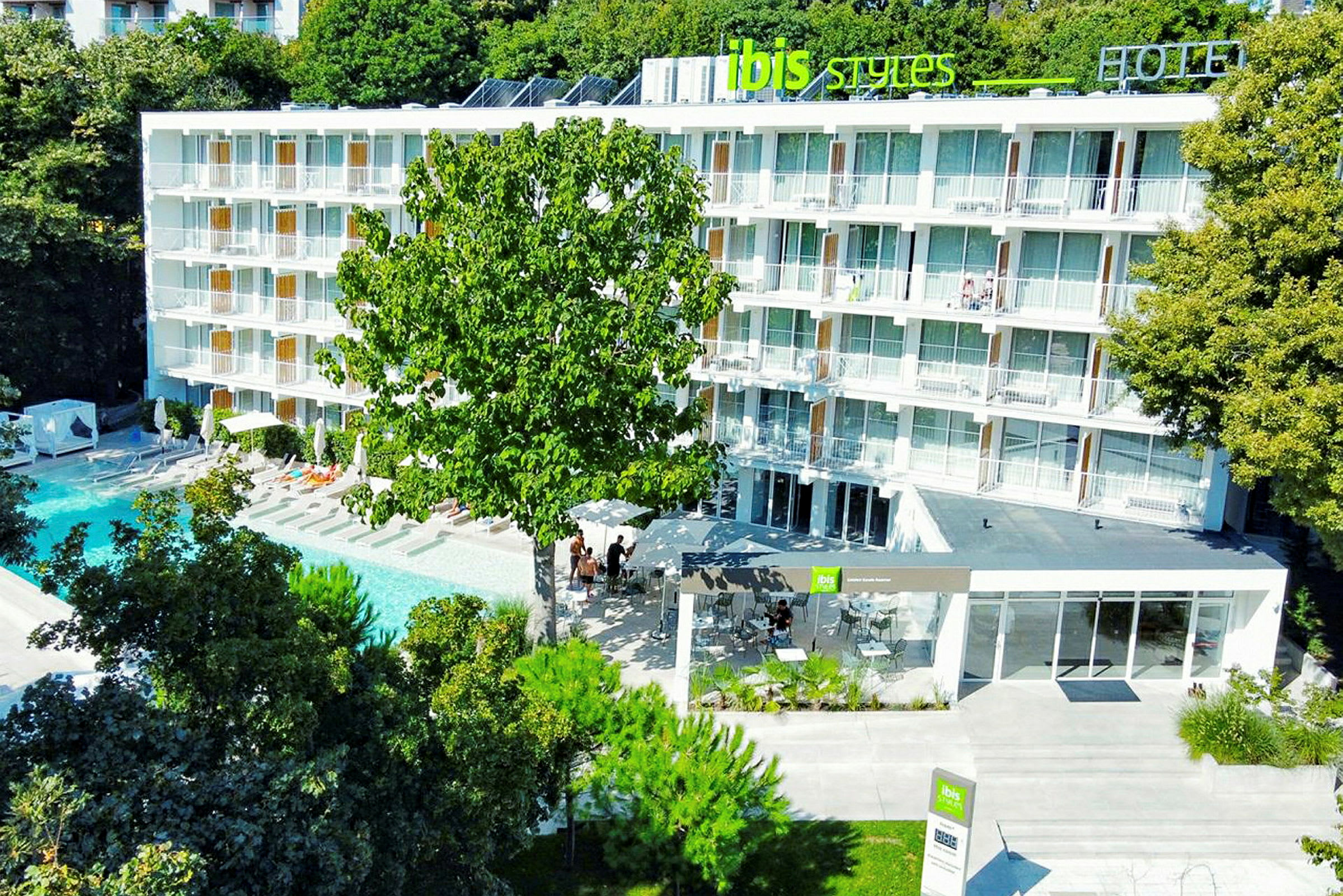Hotelbygning med altaner foran en swimmingpool, omgivet af træer. (AI-genereret tekst)