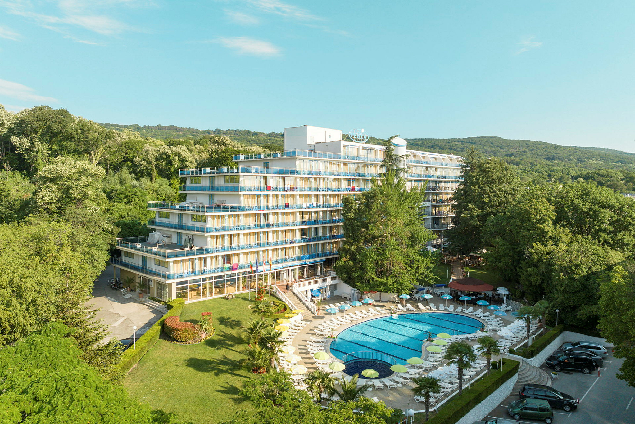 Billede af et hotel med flere etager og altaner, omgivet af frodigt grønt. En pool med liggestole ligger i forgrunden. (AI-genereret tekst)