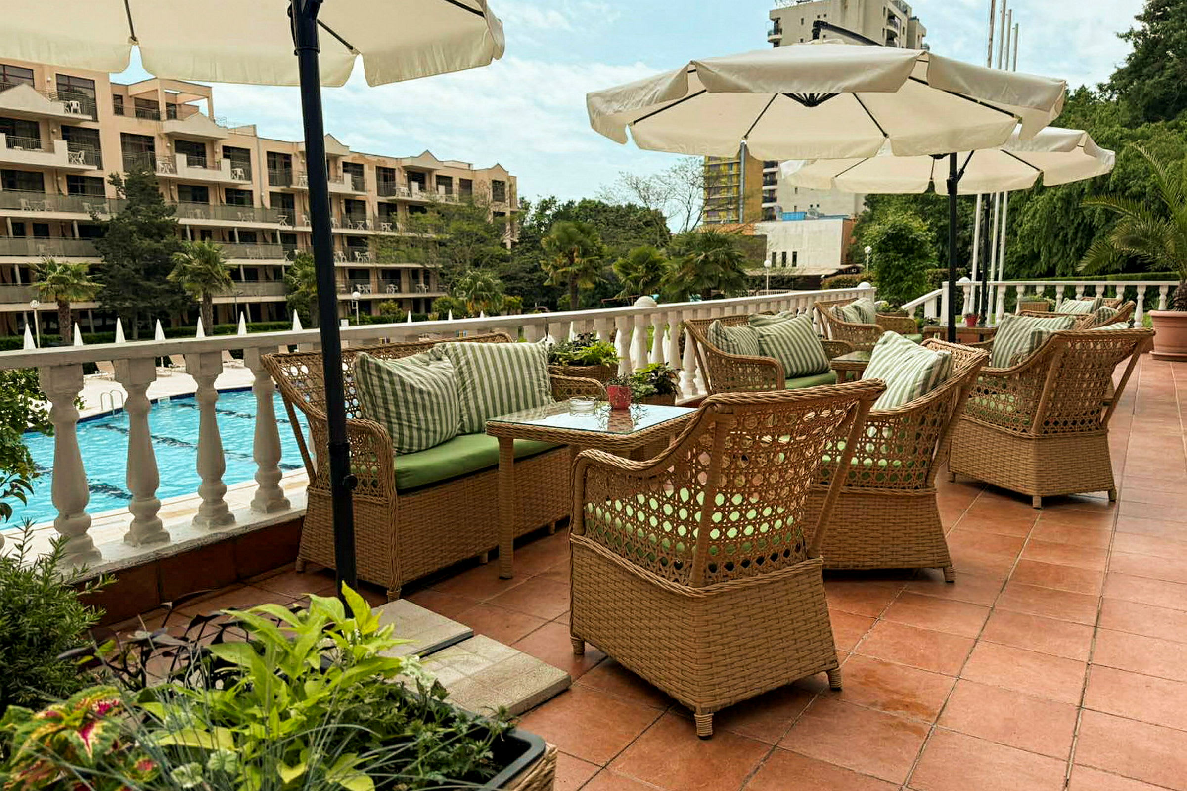 En terrasse med rottingmøbler og parasoller, med udsigt over en pool og omkringliggende bygninger. (AI-genereret tekst)