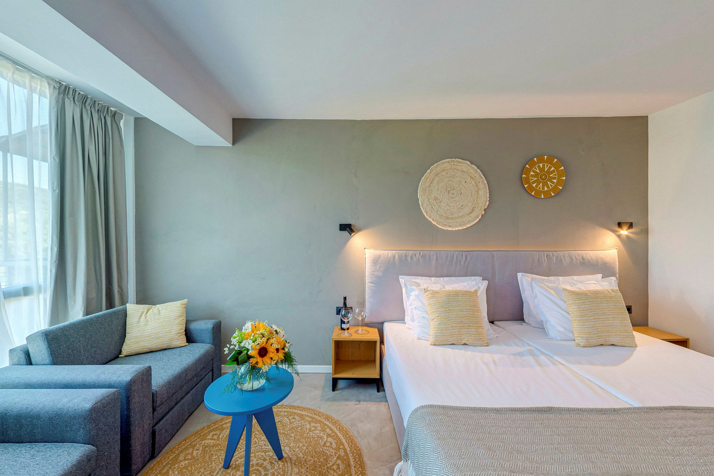 Et lyst indrettet hotelværelse med en dobbeltseng, sofa og et lille bord. På bordet står en vase med blomster. (AI-genereret tekst)