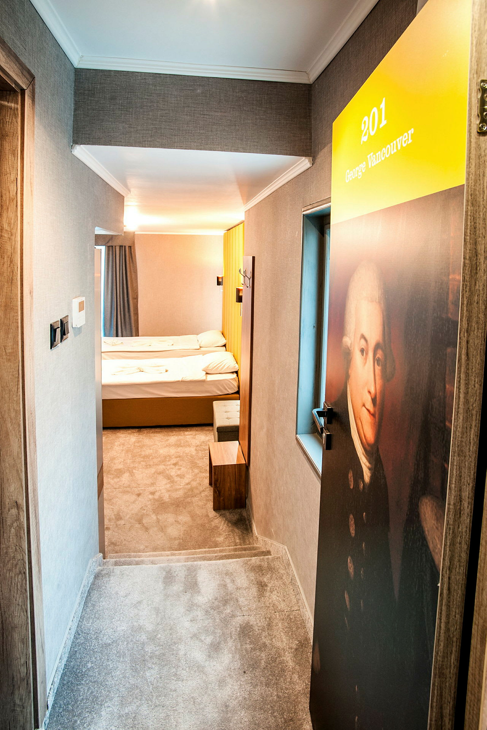 En hotelgang med en dør markeret med nummer 201 og navnet George Vancouver. Indenfor ses et enkelt hotelværelse med to senge. (AI-genereret tekst)
