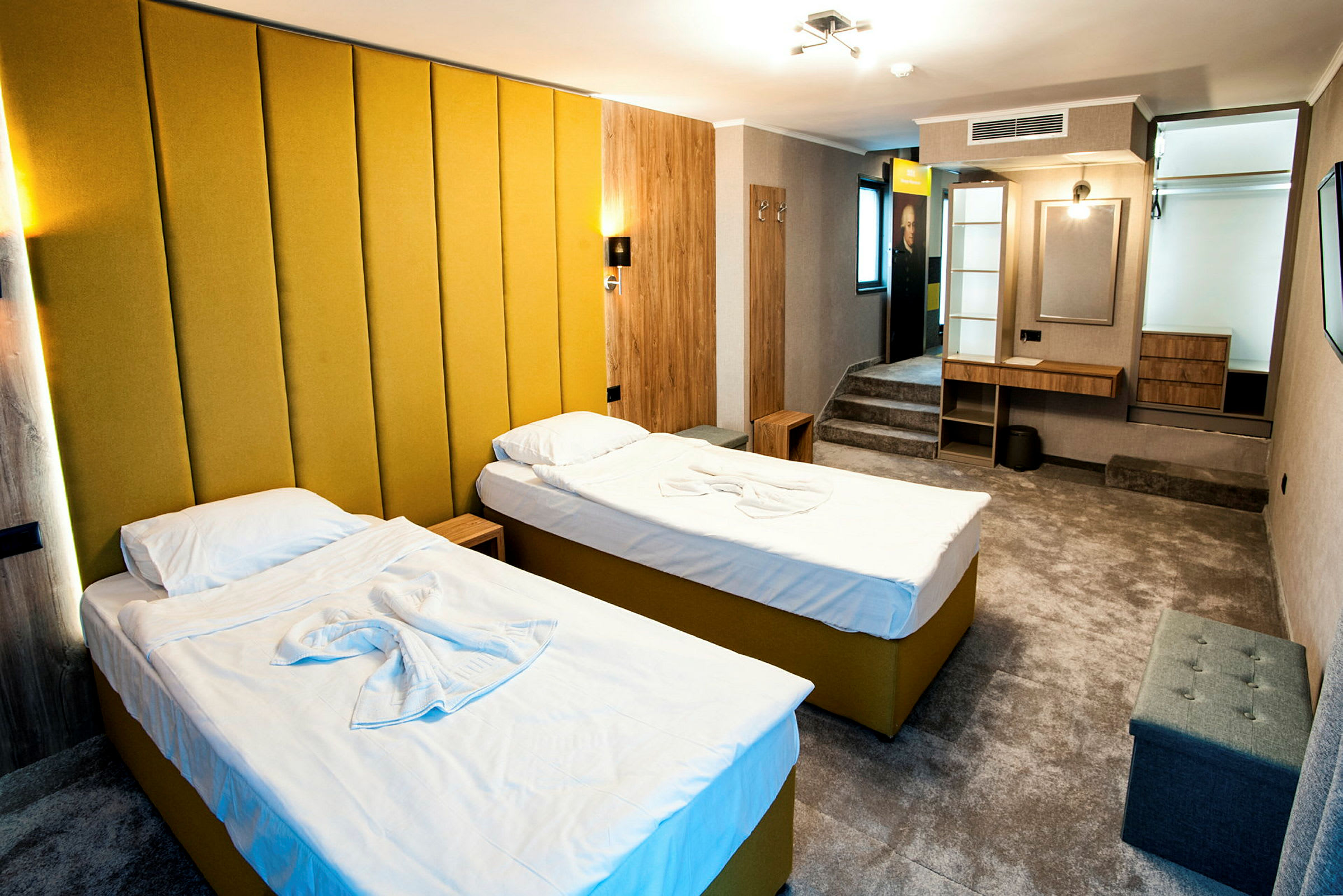 Et hotelværelse med to enkeltsenge med gul polstring og hvidt sengetøj. Værelset har en moderne indretning med en gul vægpanel, et skrivebord, siddepladser og en lille opbevaringshylde. (AI-genereret tekst)