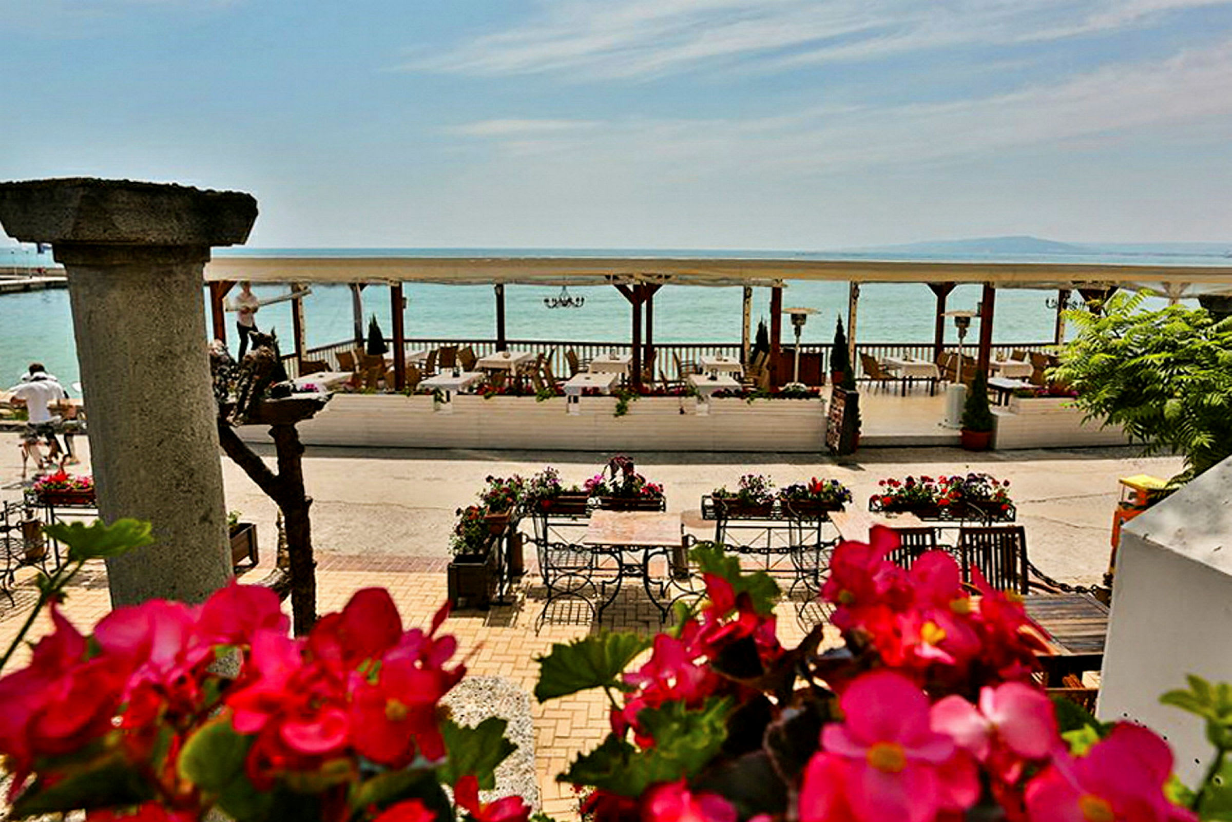 En strandnær terrasse med borde og stole omgivet af blomster og udsigt over havet. (AI-genereret tekst)