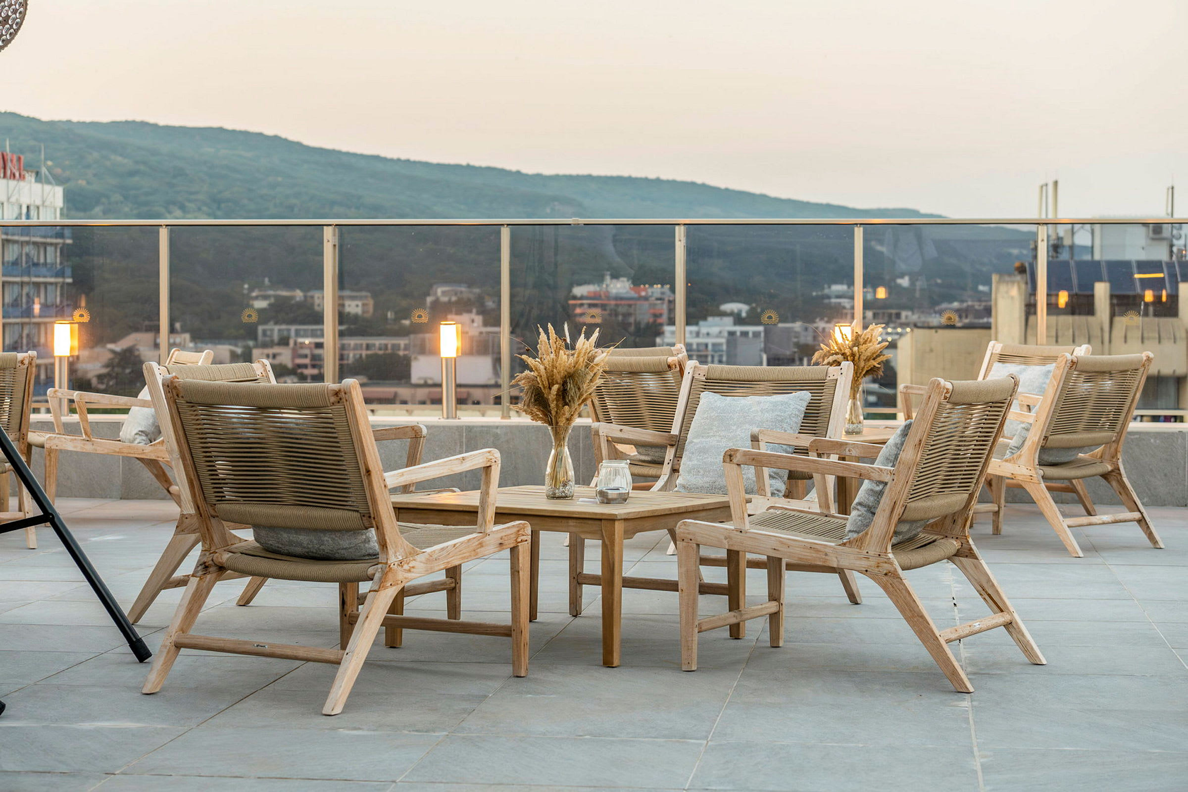 Terrasse med en gruppe træmøbler, herunder stole og bord. Udsigt over byen og grønne bakker i baggrunden. (AI-genereret tekst)