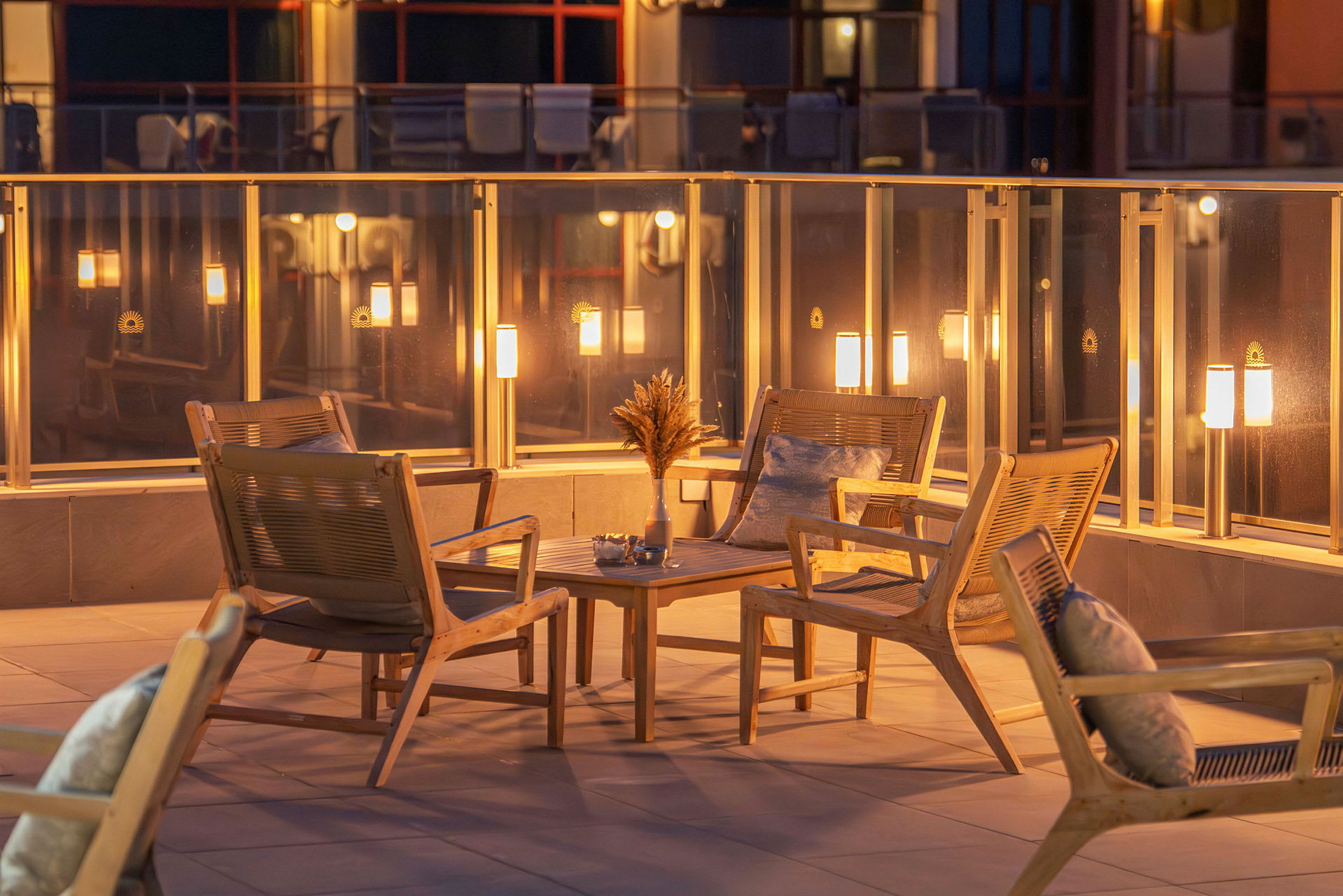 Fire træstole med puder er arrangeret omkring et lille bord på en terrasse med glasrækværk og belysning. (AI-genereret tekst)