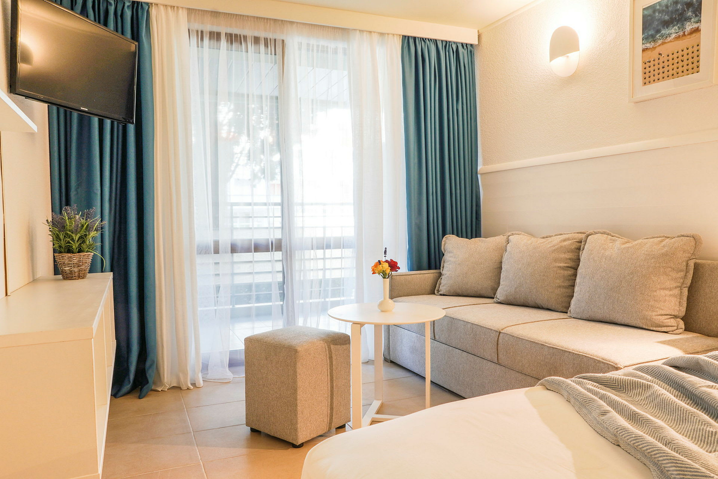 Et hotelværelse med sofa, bord, puff og TV samt et stort vindue med gardiner. (AI-genereret tekst)
