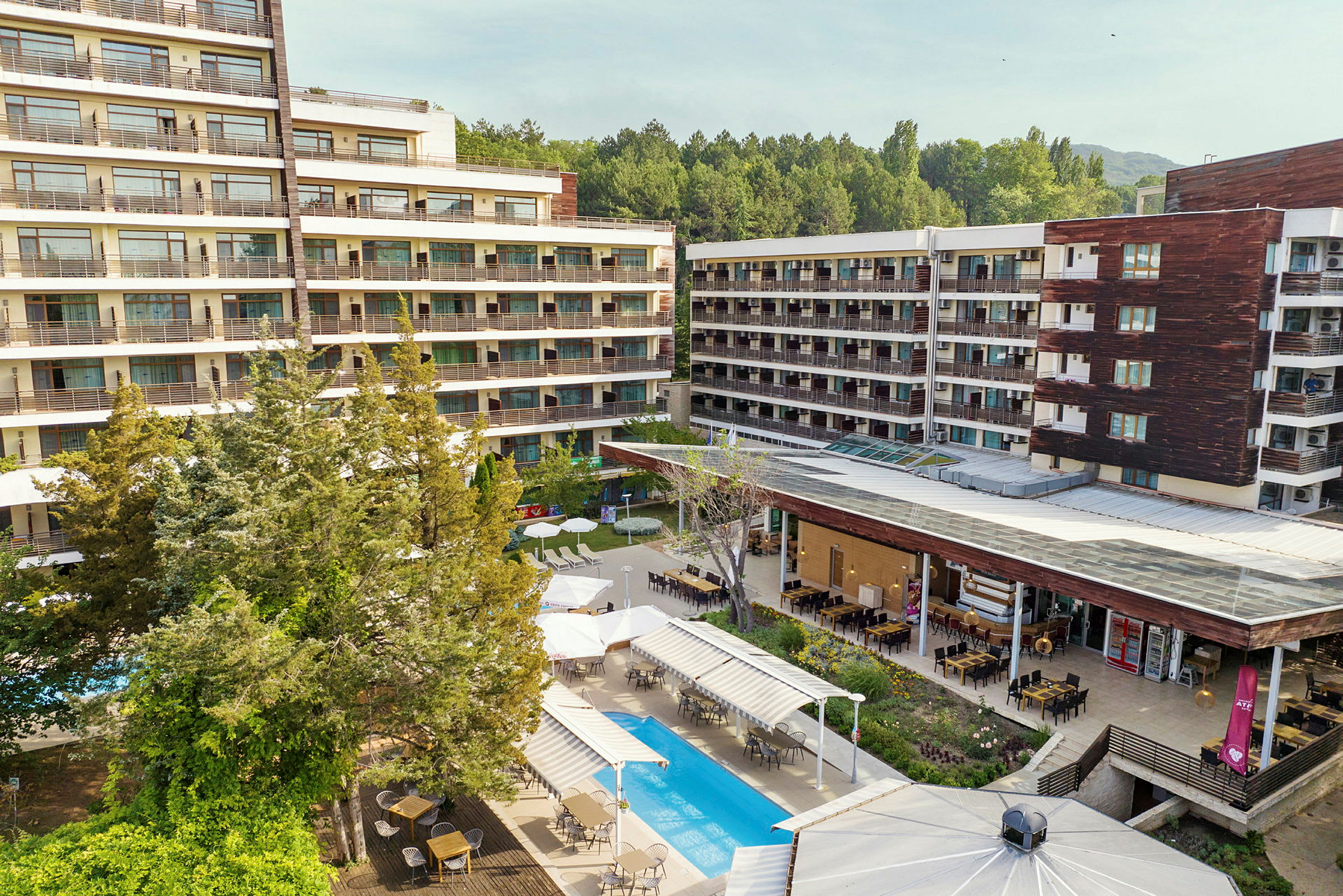 Hotelbygningskompleks omgivet af grønne områder. En pool og udendørs spiseområde ses i forgrunden. (AI-genereret tekst)