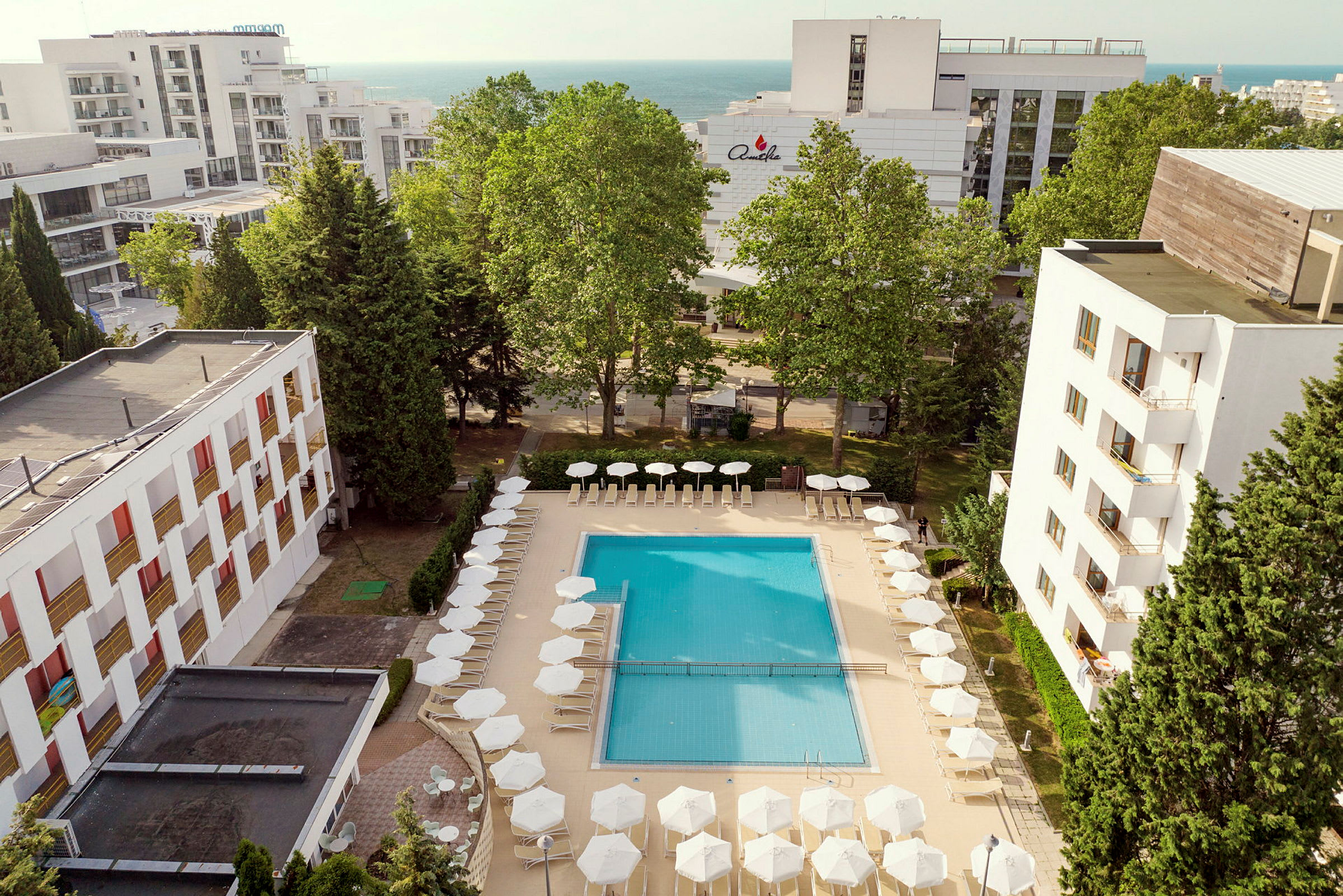 En pool omgivet af liggestole og parasoller, med hotelbygninger og træer i baggrunden. (AI-genereret tekst)