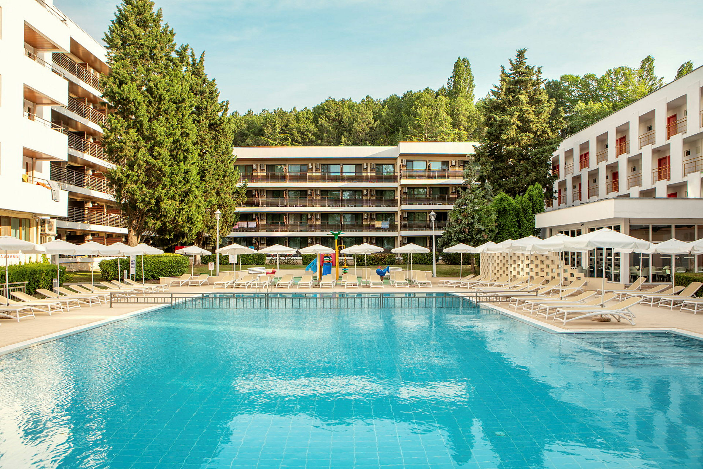 Udendørs swimmingpool med liggestole og parasoller, omgivet af moderne bygninger og grønt. (AI-genereret tekst)