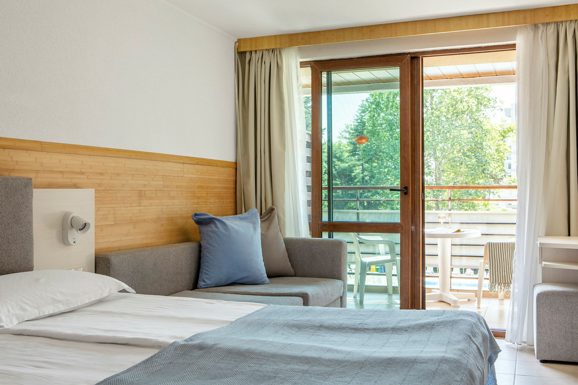 Et hotelværelse med en seng, sofa og altanmøbler synlige gennem en glasdør. (AI-genereret tekst)