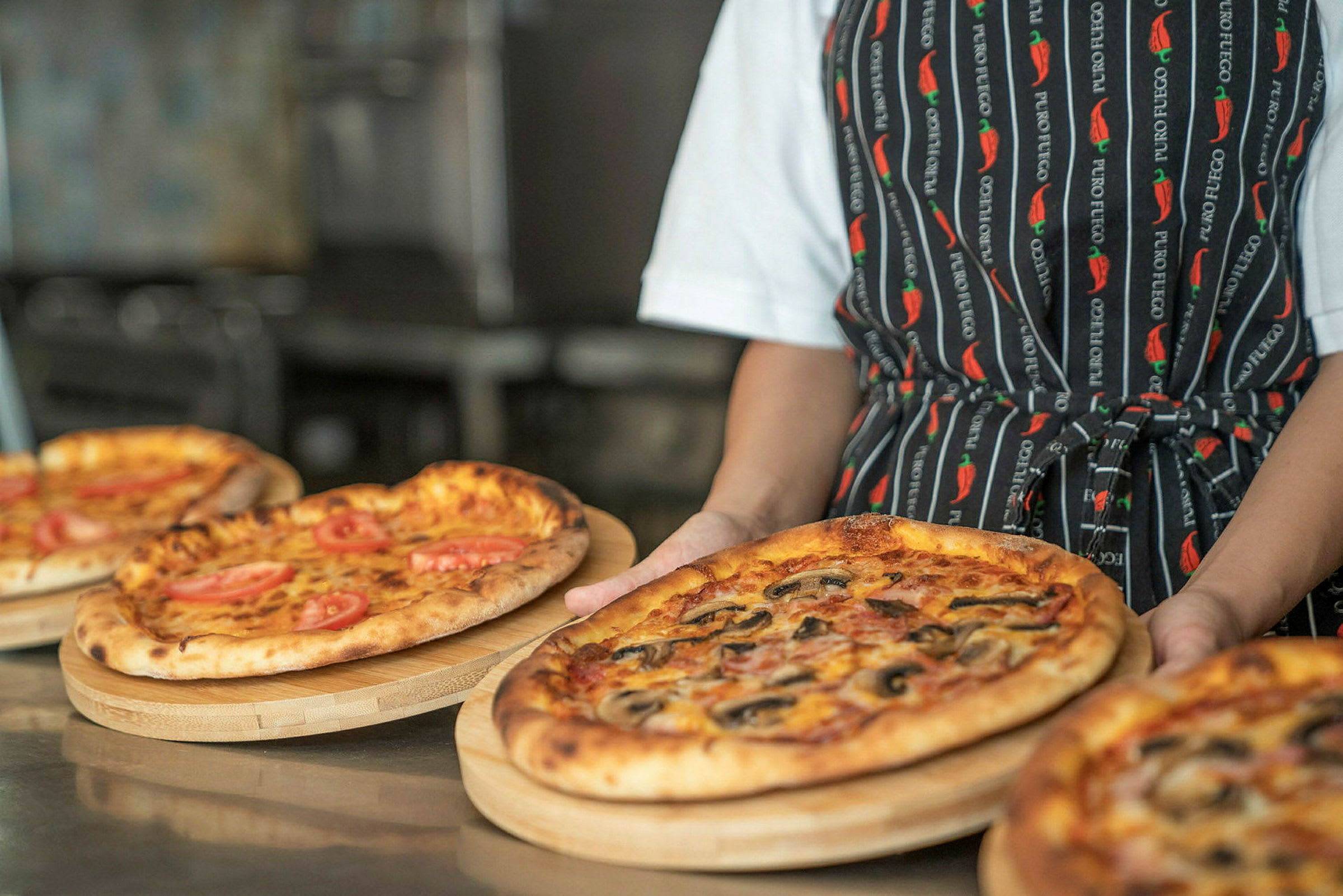 En person holder en træbakke med en pizza, omgivet af andre pizzaer. (AI-genereret tekst)
