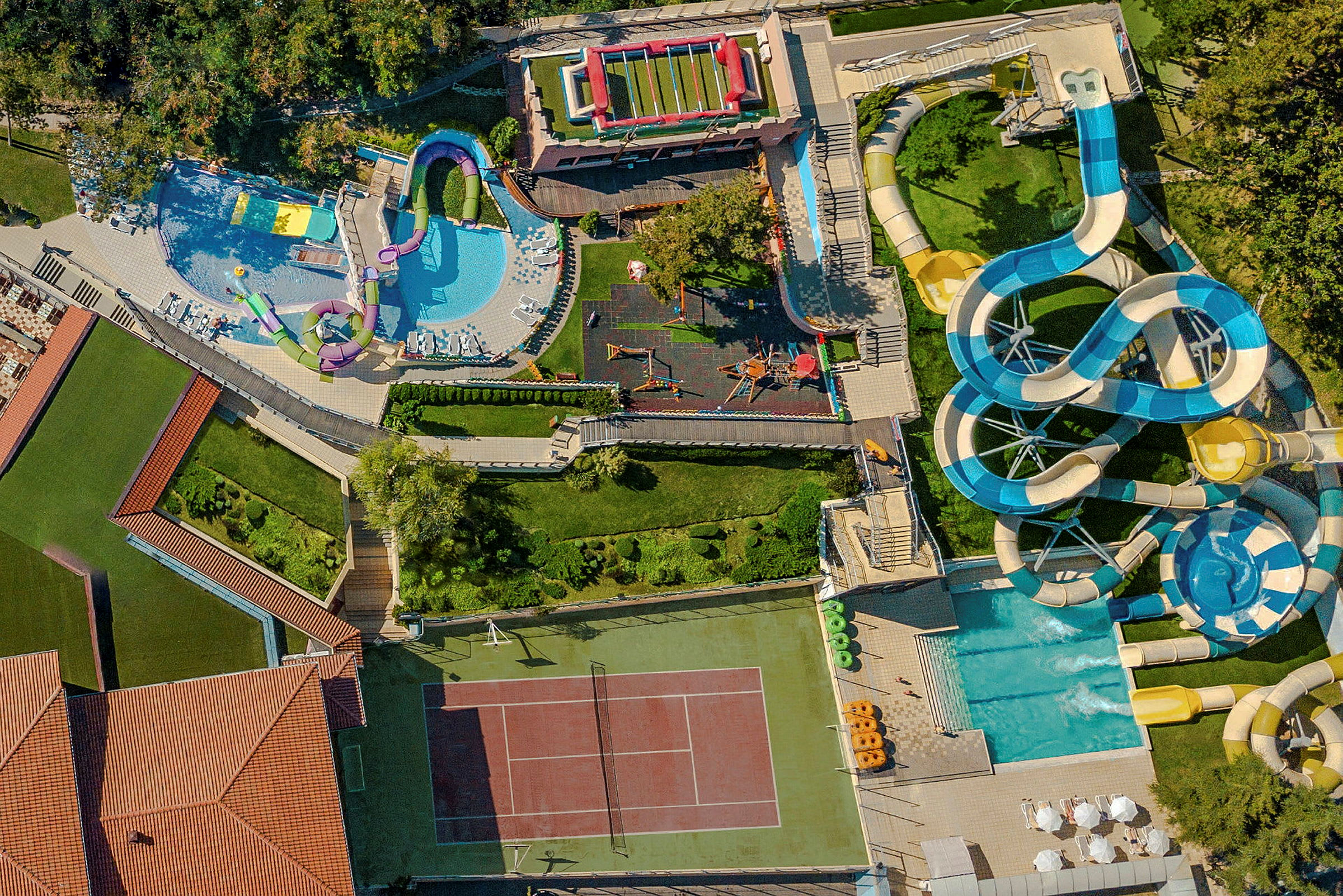 Luftfoto af et anlæg med en tennisbane, swimmingpools, vandrutsjebaner og legepladser, omgivet af grønne områder. (AI-genereret tekst)