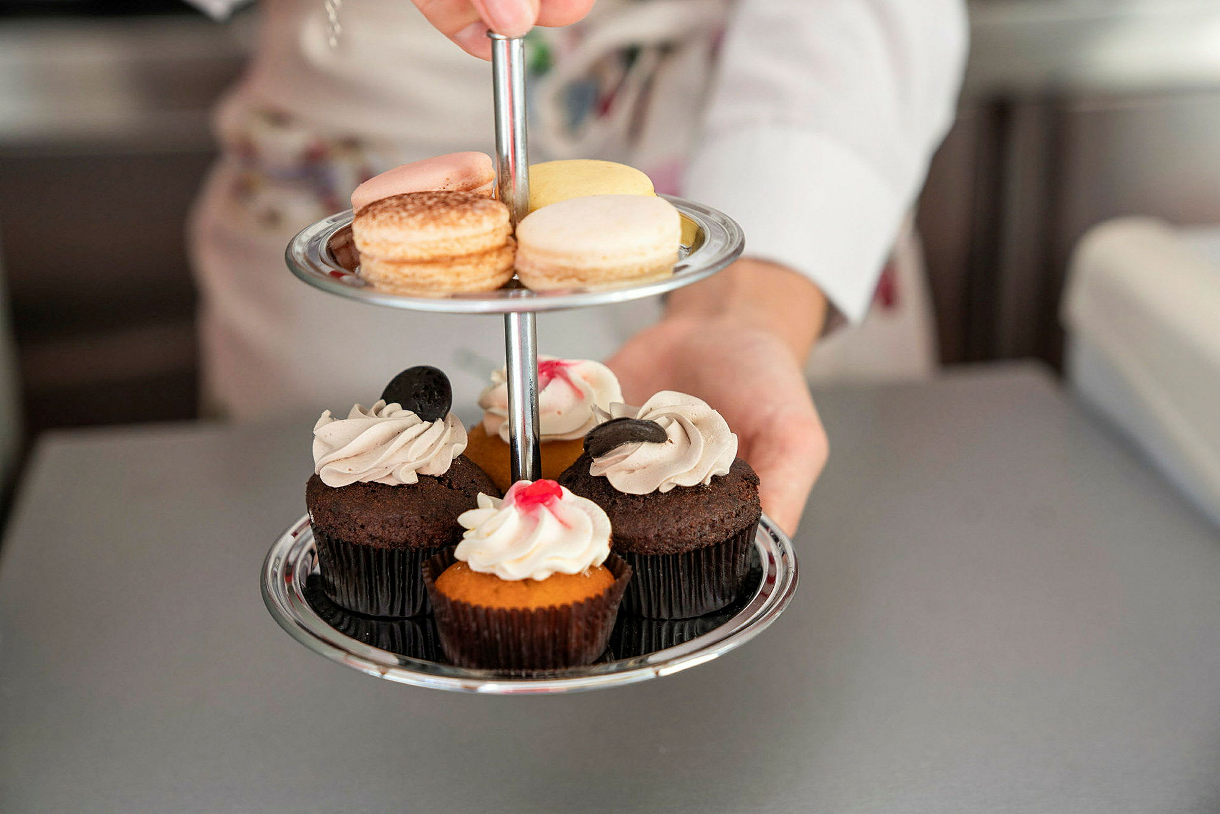 En person holder et to-etagers serveringsfad med macarons og muffins. (AI-genereret tekst)