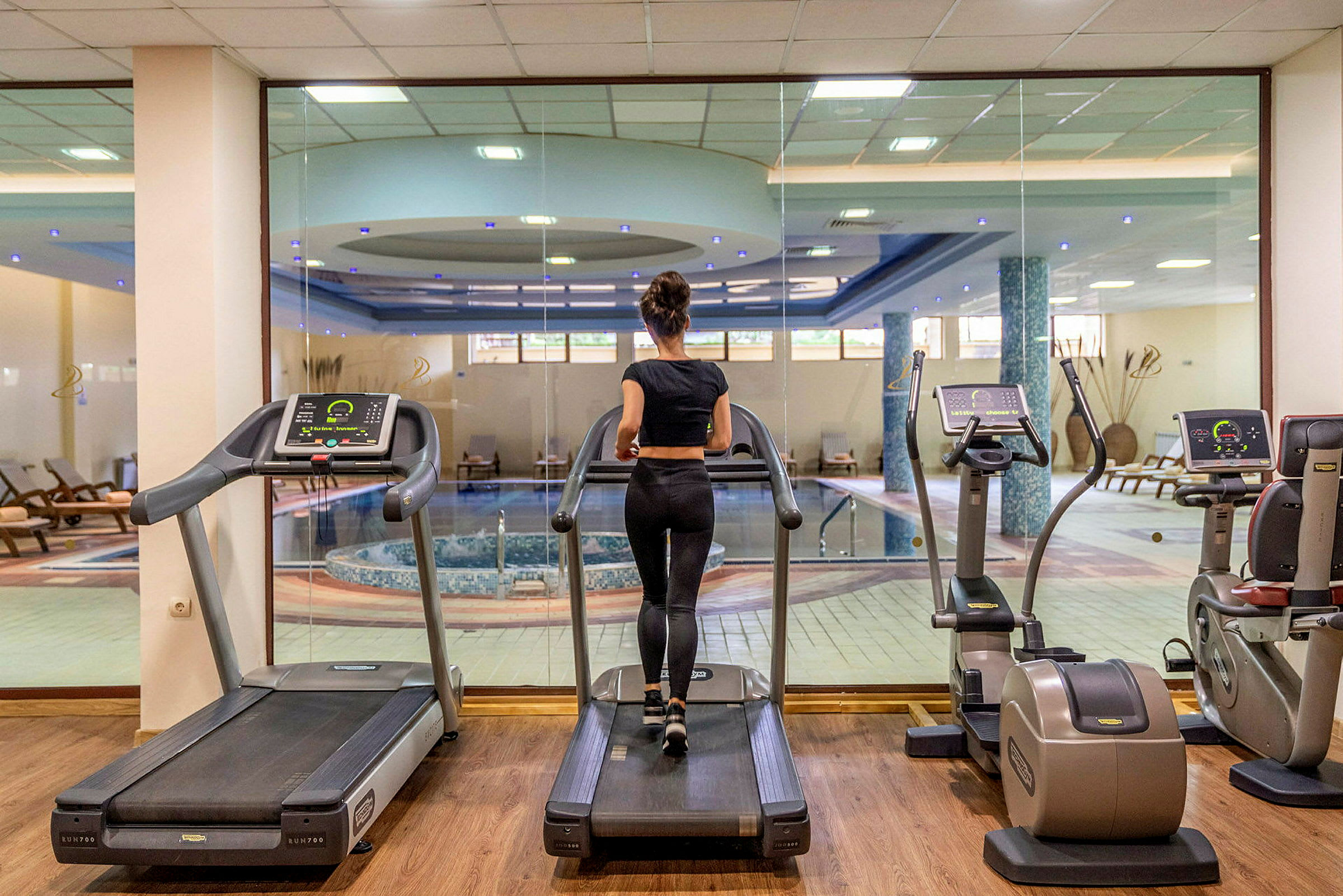 Kvinde træner på et løbebånd i et fitnesscenter med udsigt til en indendørs pool. (AI-genereret tekst)