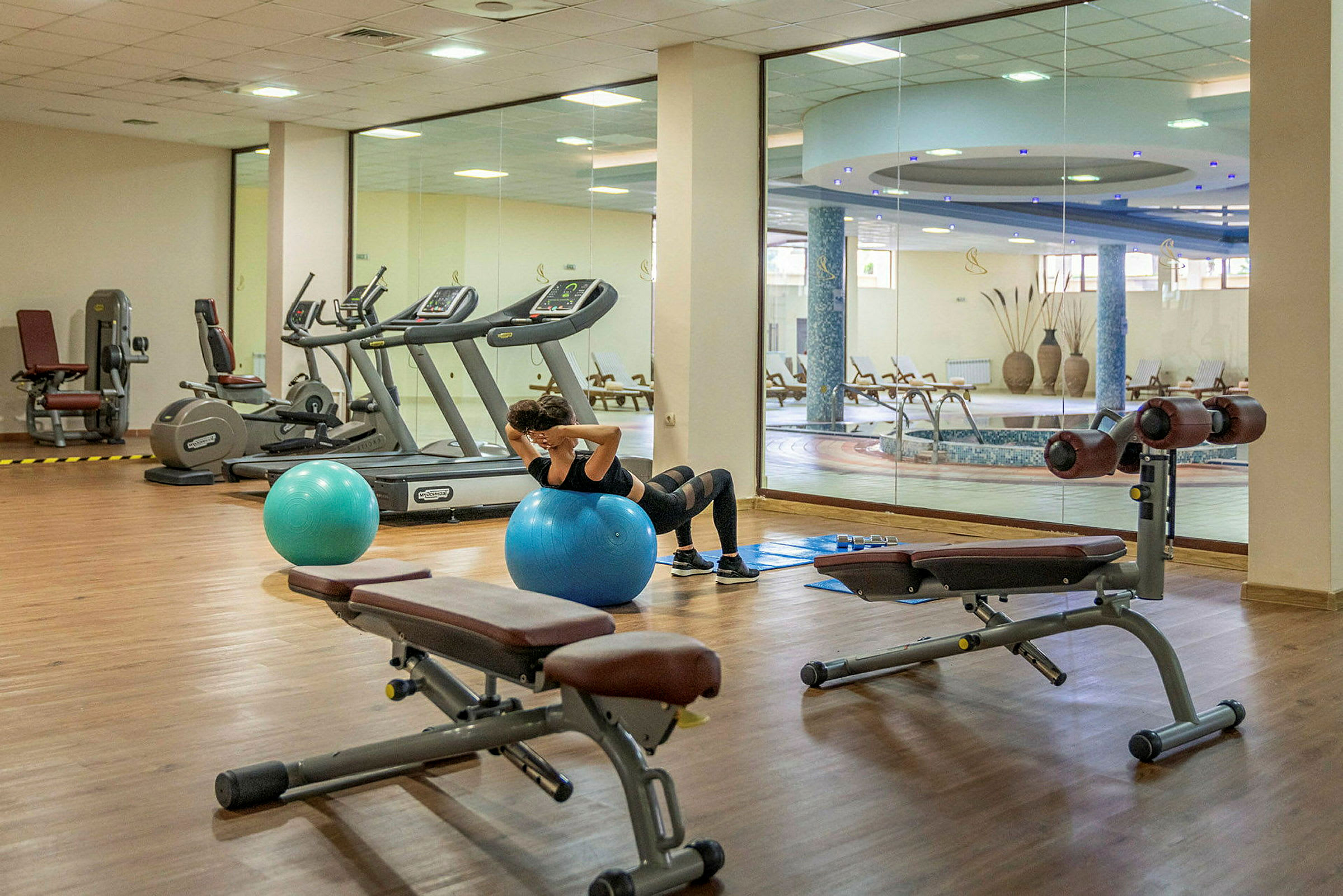 En person træner på en blå træningsbold i et fitnesscenter med fitnessudstyr og udsigt til en indendørs pool. (AI-genereret tekst)