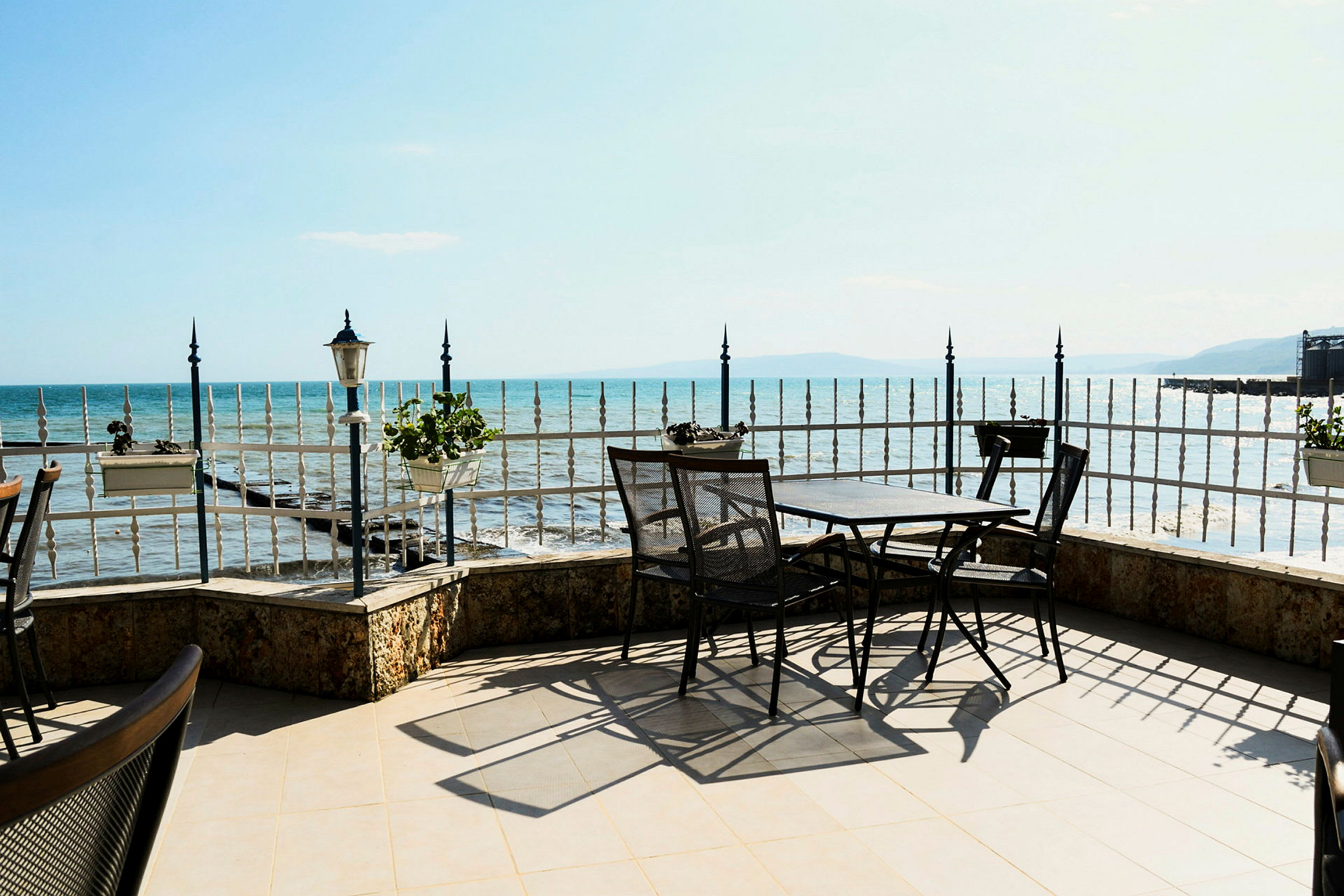 Terrasse med bord og stole ved et jerngitter, med udsigt over havet i klart vejr. (AI-genereret tekst)