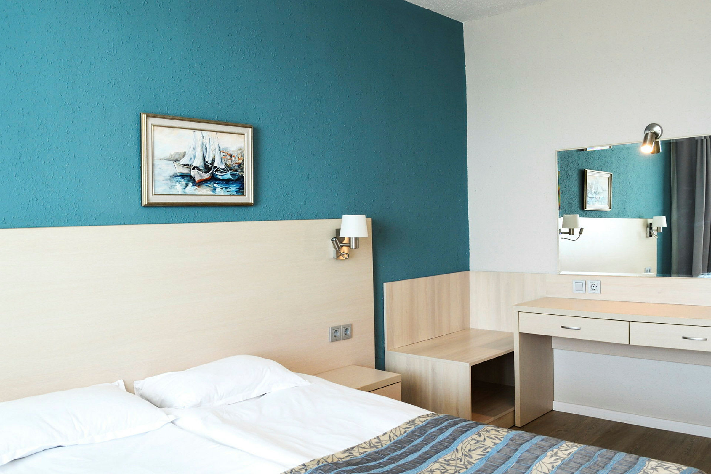Hotelværelse med dobbeltseng, blå væg, maleri, senge lamper og skrivebord med spejl. (AI-genereret tekst)