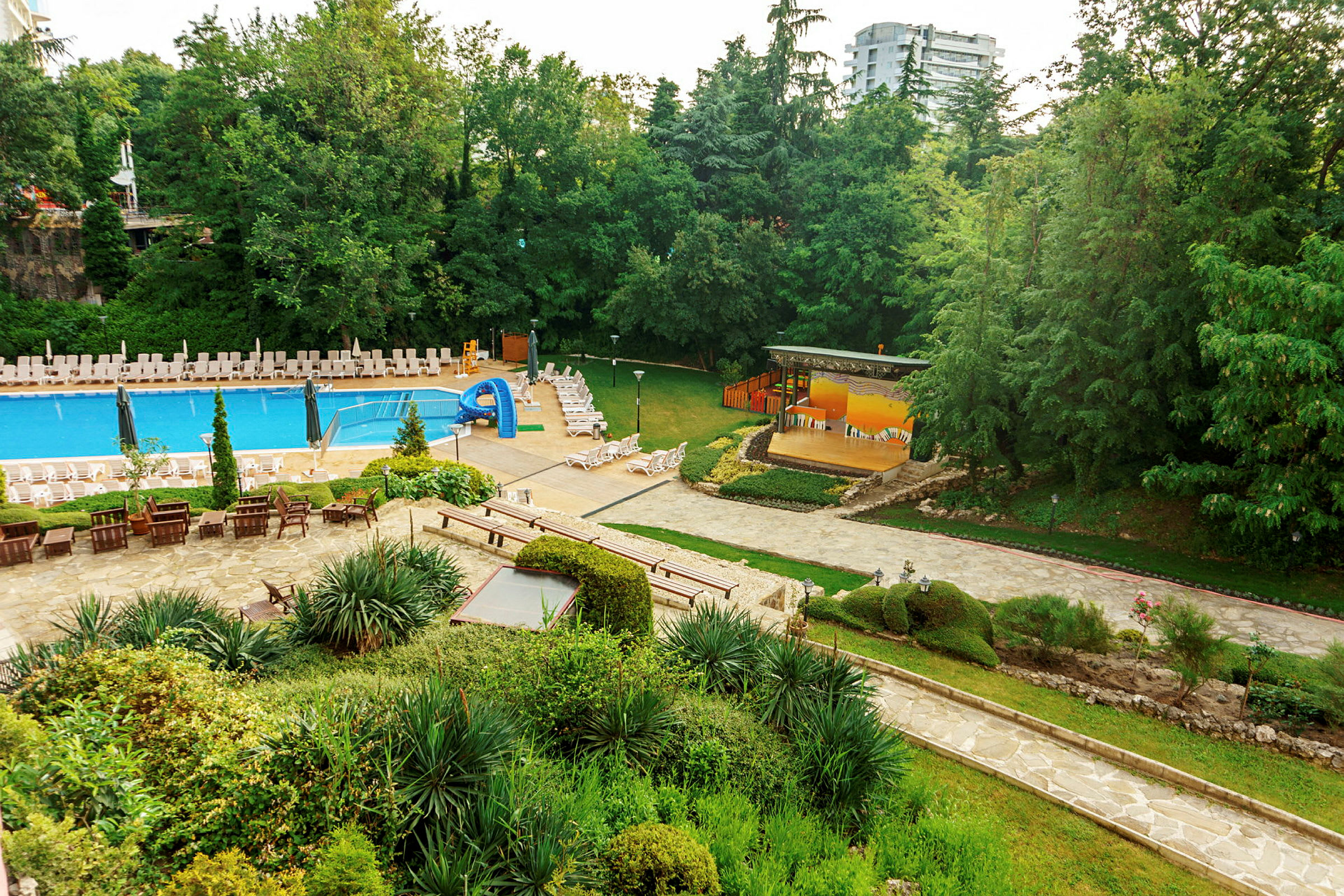 Udendørsområde med pool, liggestole, grønne planter og et sceneområde. (AI-genereret tekst)