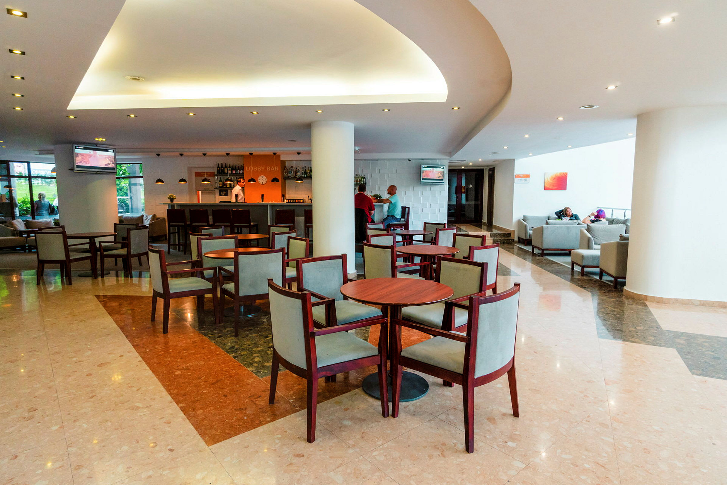 Billede af et hotellobbyområde med borde og stole, en bar i baggrunden og siddegrupper på siden. (AI-genereret tekst)