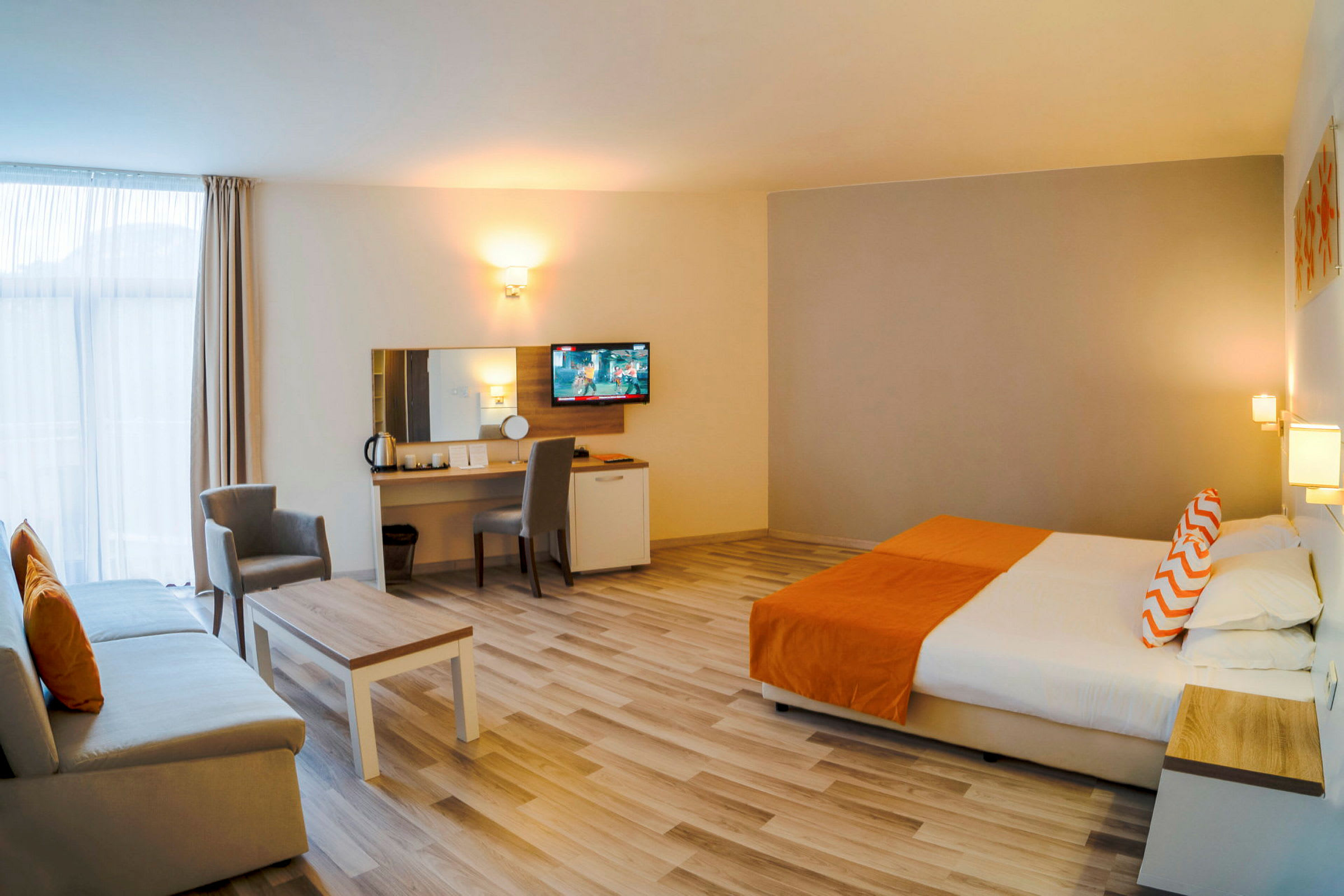 Billede af et rummeligt hotelværelse med en stor seng, sofa, bord og arbejdsområde, indrettet i neutrale og orange farver. (AI-genereret tekst)
