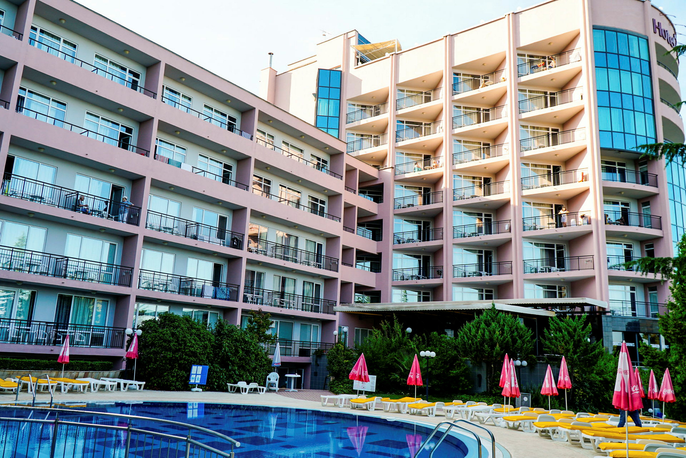 Et større hotel med flere etager og altaner med udsigt til en pool samt liggestole og parasoller. (AI-genereret tekst)