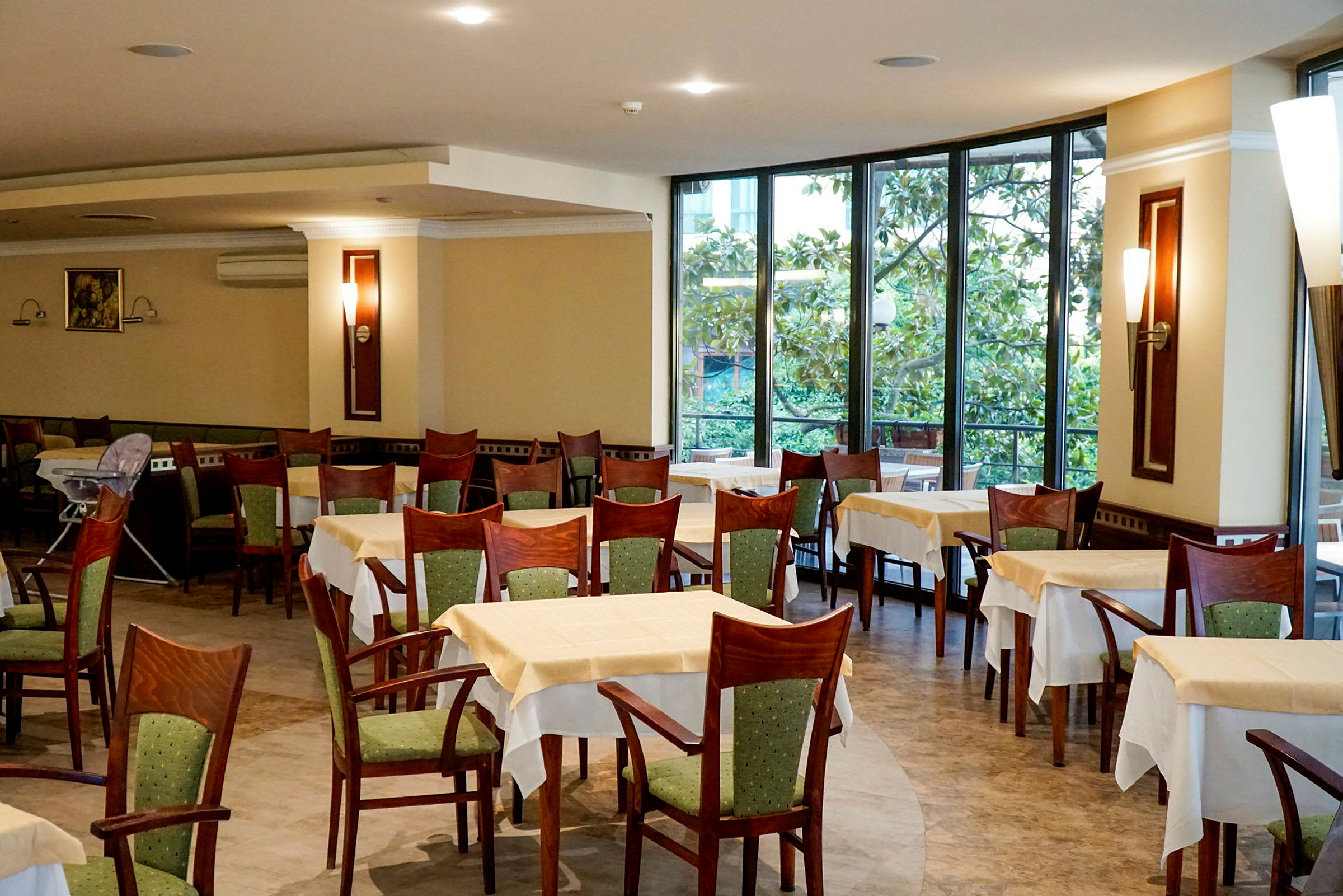 Restaurant med borde og stole, store vinduer og grønne træer udenfor. (AI-genereret tekst)