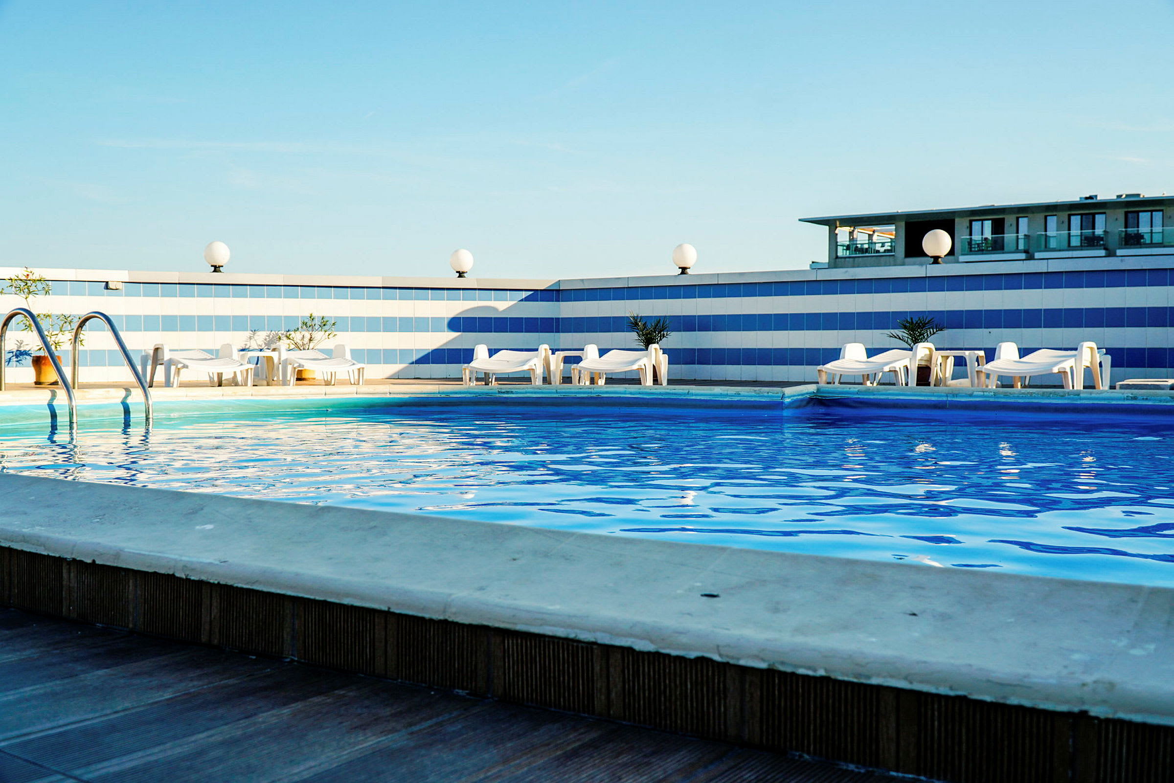 En udendørs pool omgivet af liggestole med blå himmel i baggrunden. (AI-genereret tekst)