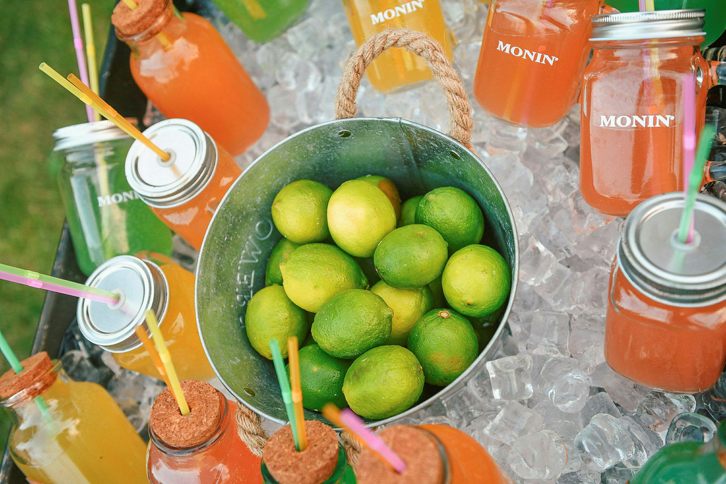 En metalspand fyldt med limefrugter omgivet af glas med forskelligfarvede drikke og isterninger. (AI-genereret tekst)