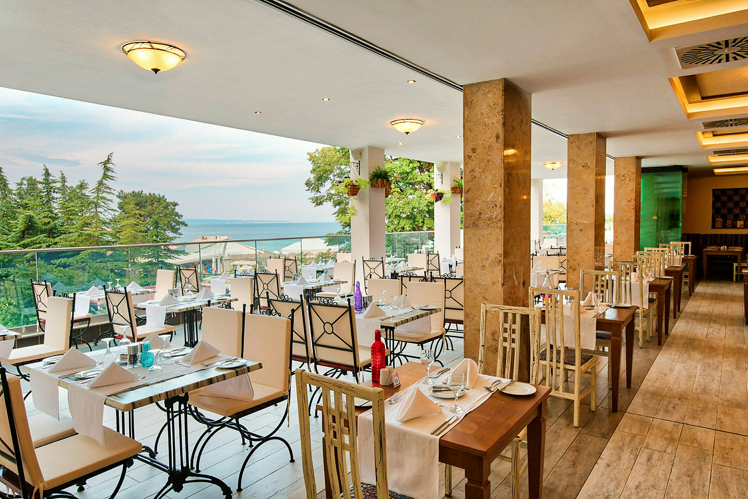 En restaurant med borde og stole i lyse farver, udsigt over havet og omgivet af træer. (AI-genereret tekst)