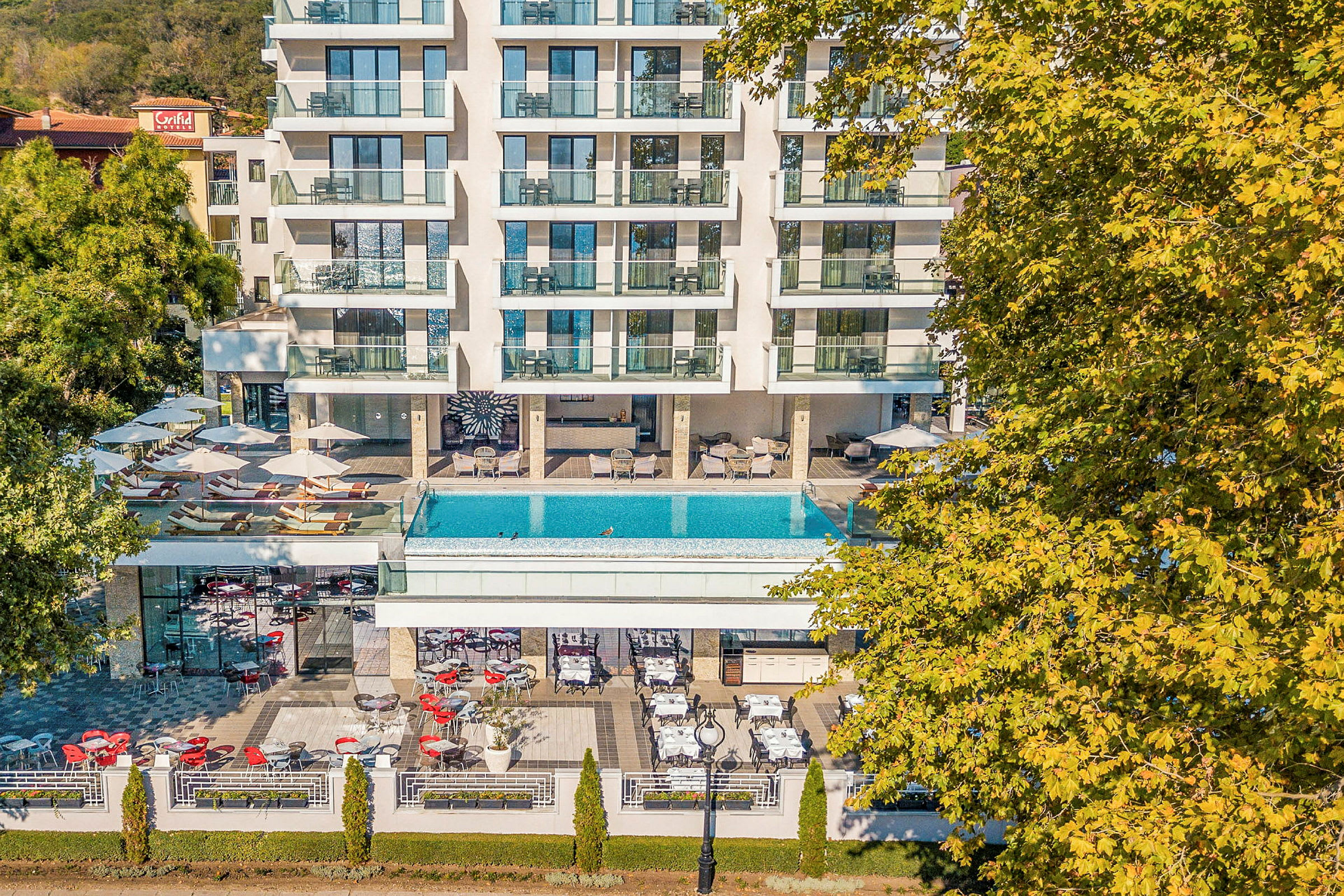 Hotel med altaner, pool og udendørsservering omgivet af grøntområder. (AI-genereret tekst)