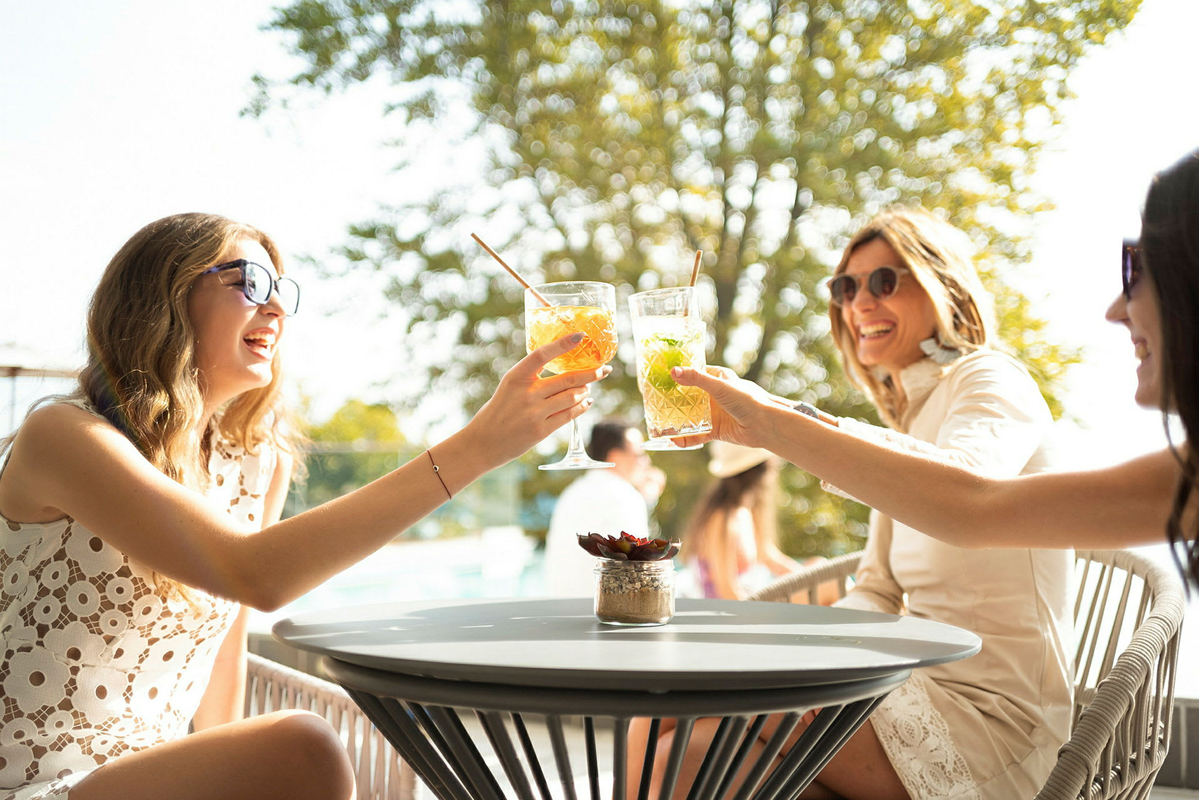 Personer, der skåler med drinks ved et bord udendørs med træer i baggrunden. (AI-genereret tekst)