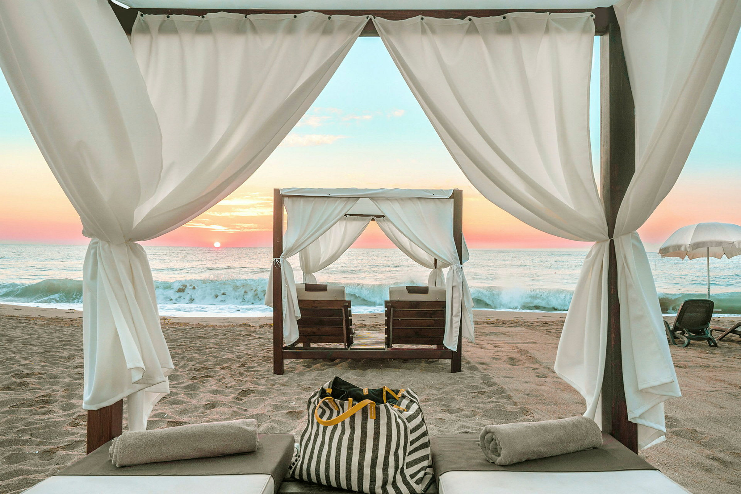 En opredt solseng under en skyggefuld cabana på en sandstrand ved havet ved solnedgang. (AI-genereret tekst)