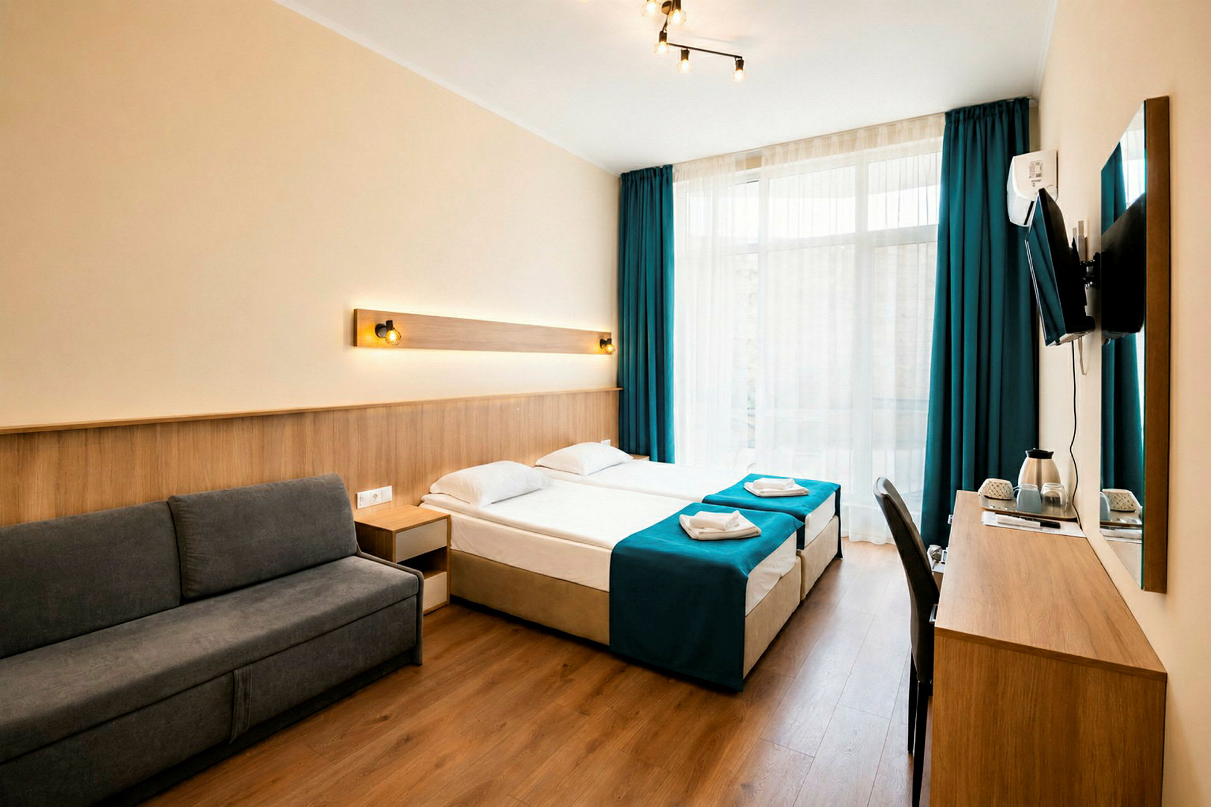 Et hotelværelse med moderne indretning, mørkegrå sofa, to enkeltsenge med blå sengetæpper og et skrivebord med et vægmonteret TV. (AI-genereret tekst)
