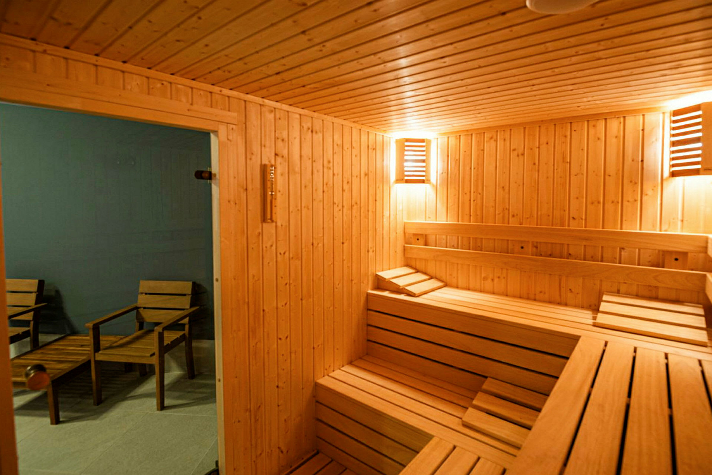 Et rum indrettet som en traditionel sauna med træpaneler og siddepladser i træ. Uden for saunaen ses stole og et bord. (AI-genereret tekst)