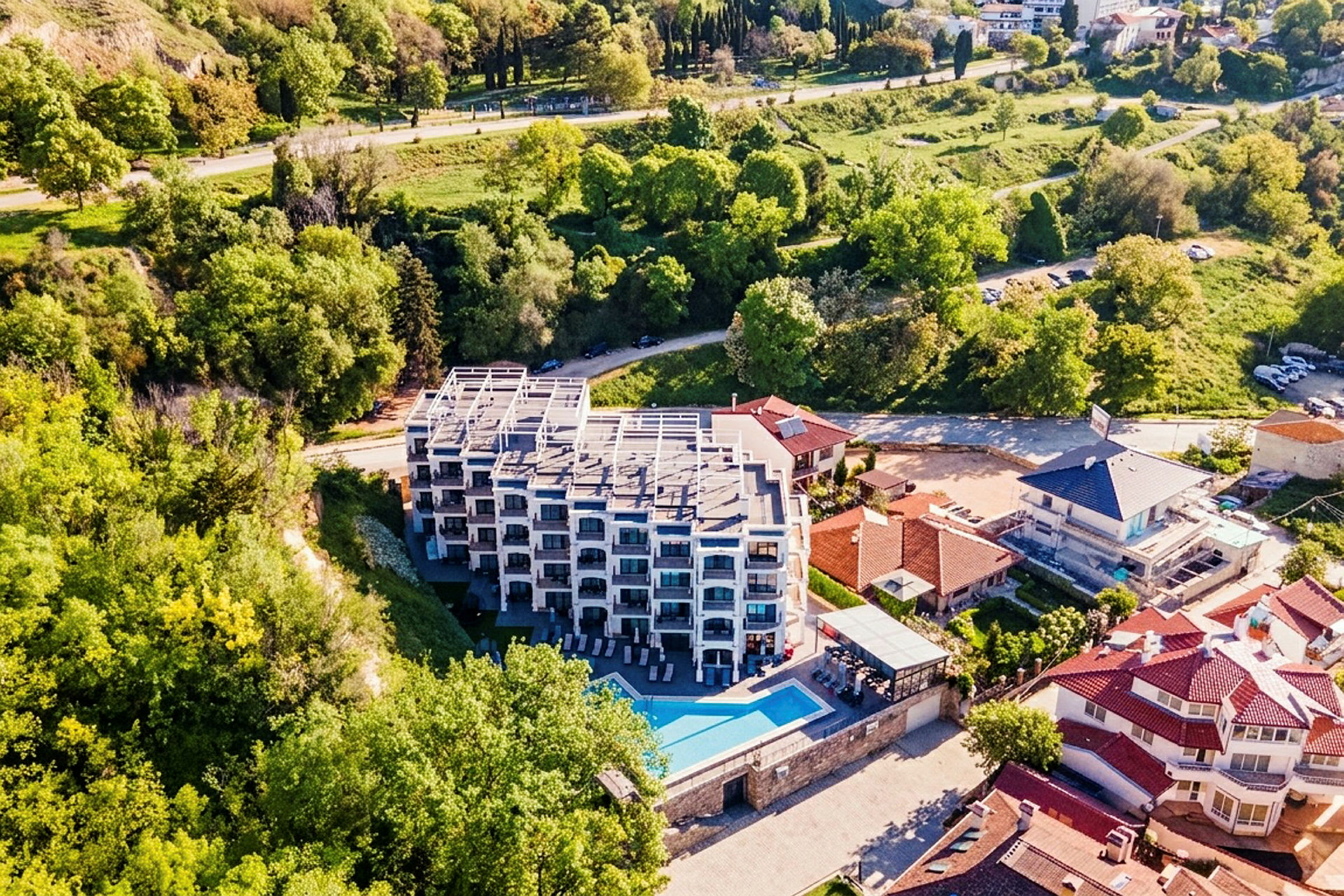 Et hotel i flere etager med altaner og en pool foran, omgivet af grønne områder og andre huse. (AI-genereret tekst)