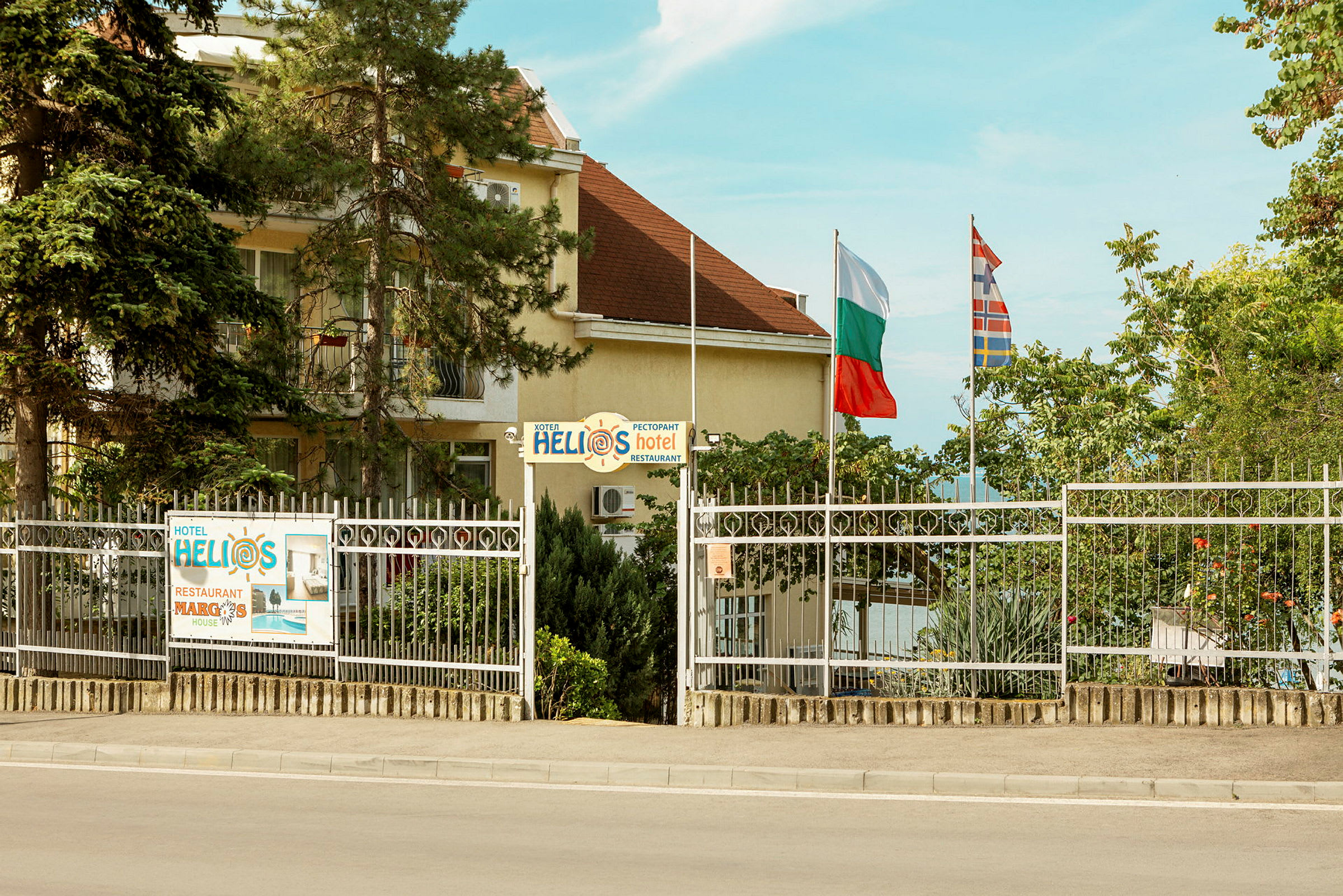Et hotel med navnet Helios, omgivet af træer og et hegn, med flag ved siden af hegnet. (AI-genereret tekst)