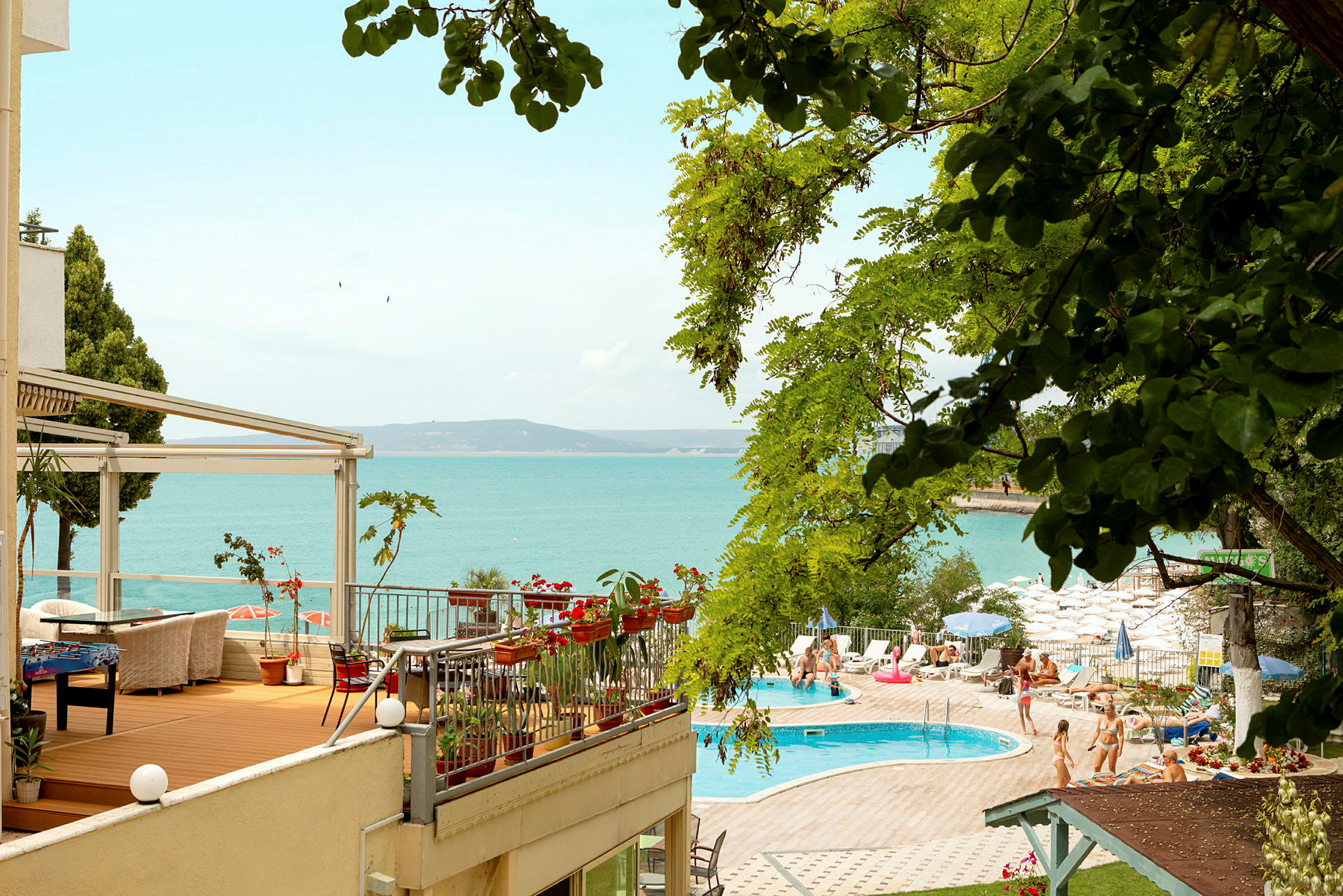 Billede af en udendørs terrasse med siddegrupper og blomster samt et poolområde og en strand ved havet. (AI-genereret tekst)