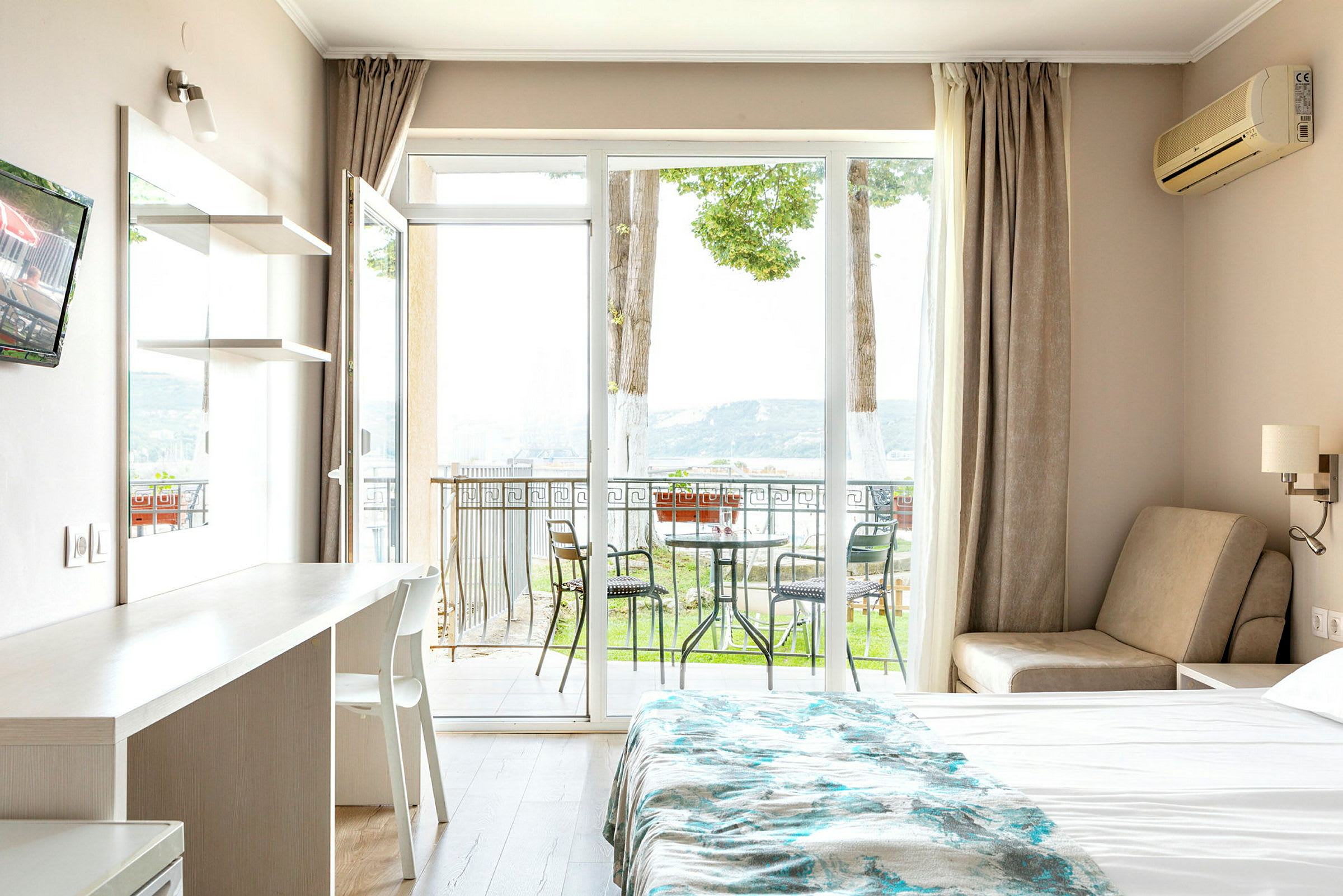 Et hotelværelse med en seng, et bord og en stol samt en balkon med havemøbler og udsigt til naturen. (AI-genereret tekst)
