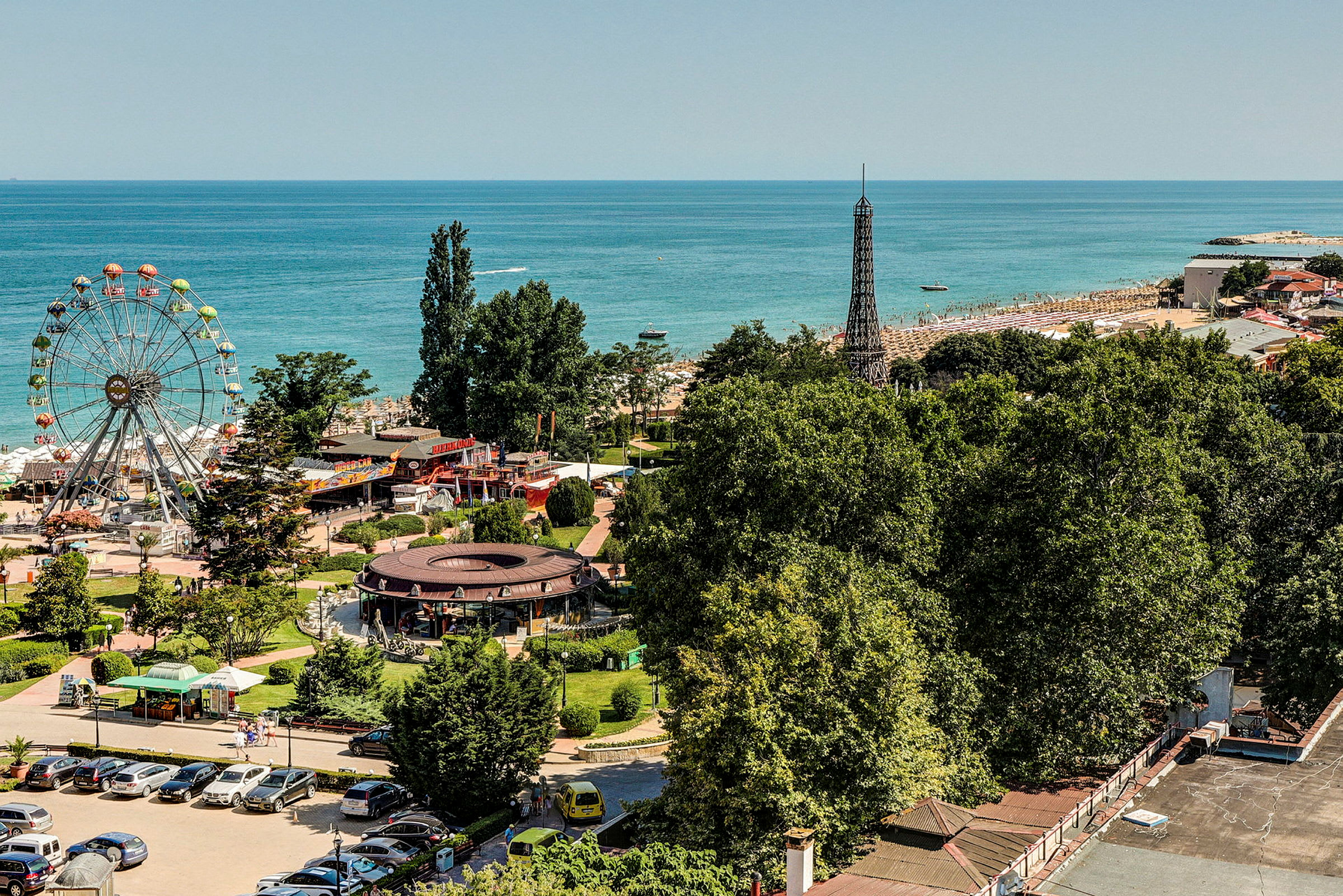 Udsigt over en forlystelsespark og en strand ved havet, med en stor pariserhjul og grønne områder. (AI-genereret tekst)