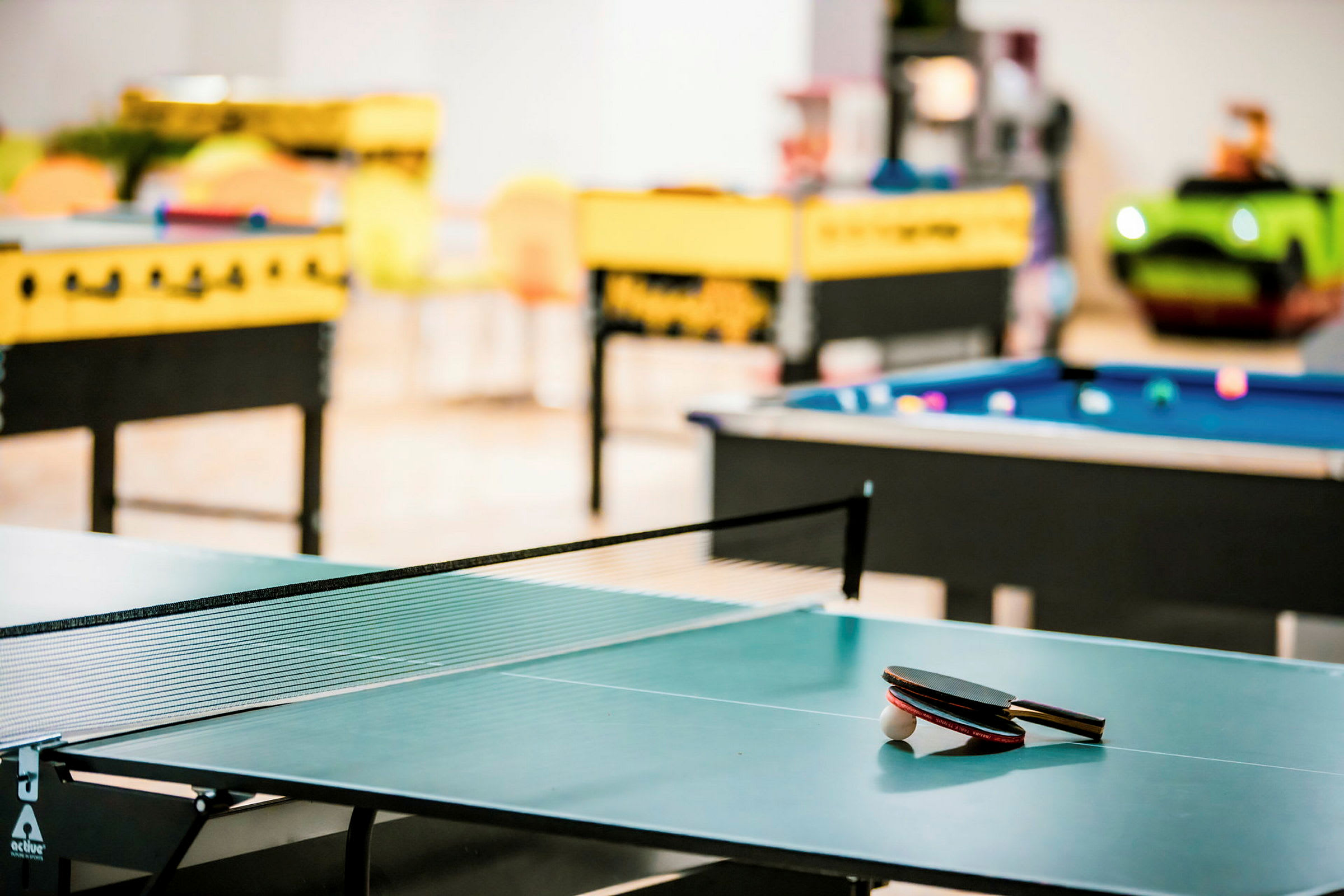 Et bordtennisbord med ketsjere og bold, med billard- og bordfodboldborde i baggrunden. (AI-genereret tekst)