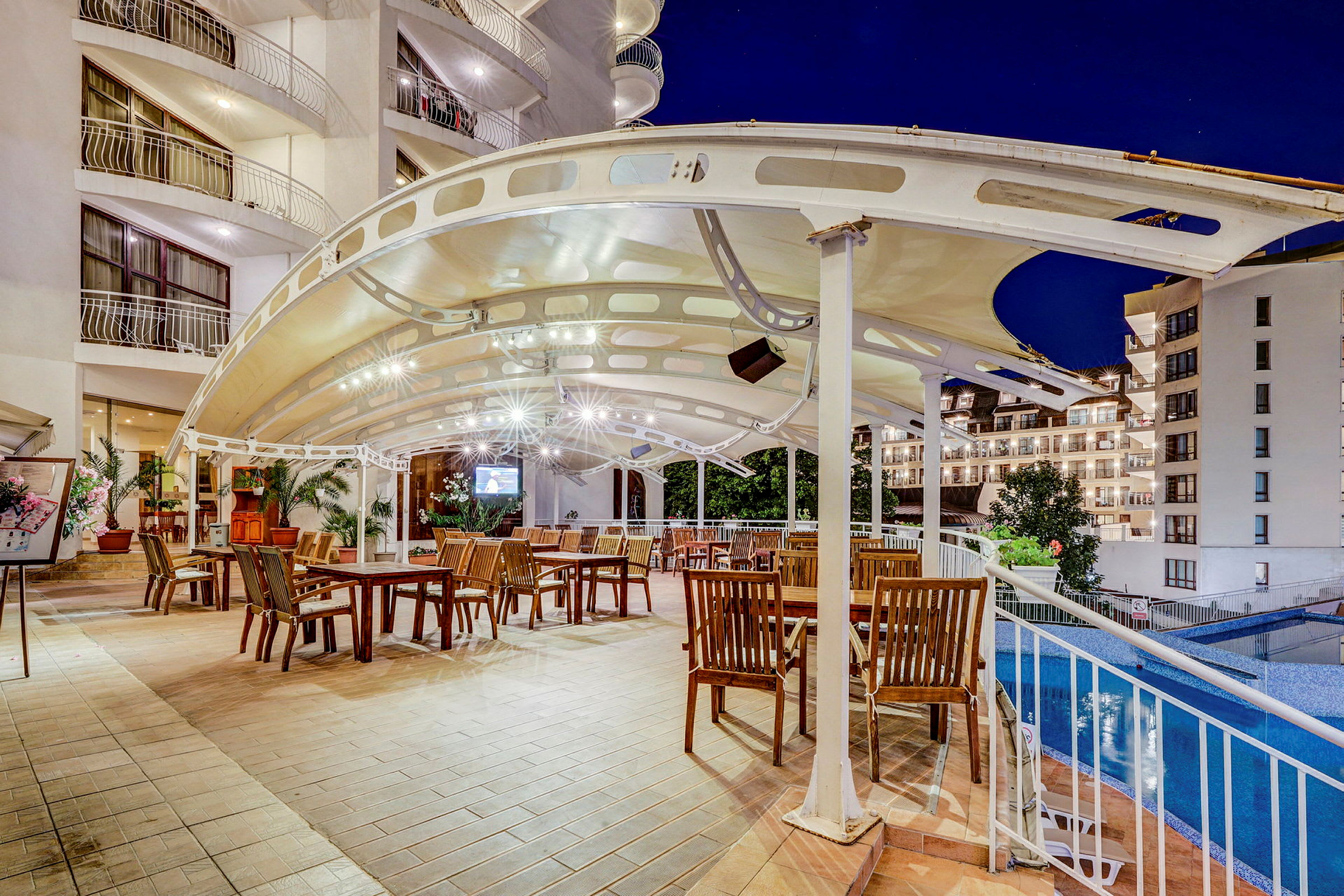 En udendørs terrasse med træborde og stole under et moderne tag, med udsigt over en pool og omkringliggende bygninger. (AI-genereret tekst)