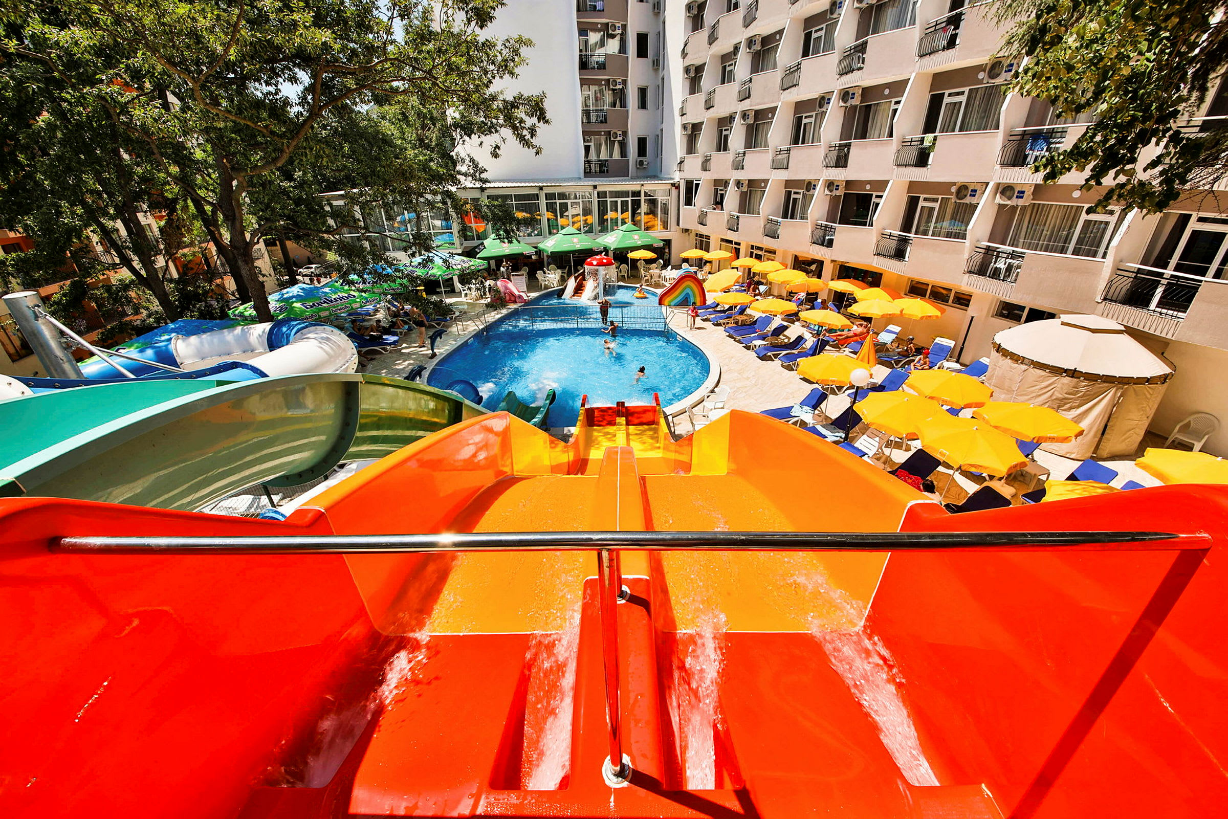 Udsigt ned fra en orange vandrutsjebane mod en udendørs pool på et hotelområde, omgivet af liggestole og parasoller. (AI-genereret tekst)