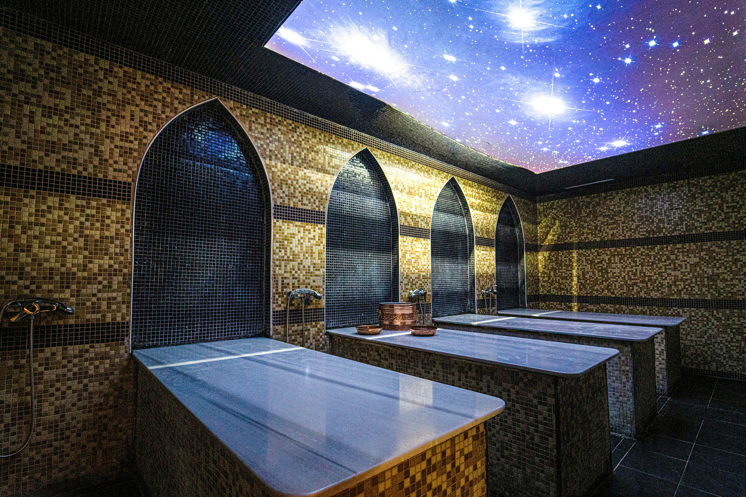Rummet viser et traditionelt hamam med marmorborde og dekorativ mosaik på væggene. Loftet er udsmykket med et motiv af stjerner. (AI-genereret tekst)