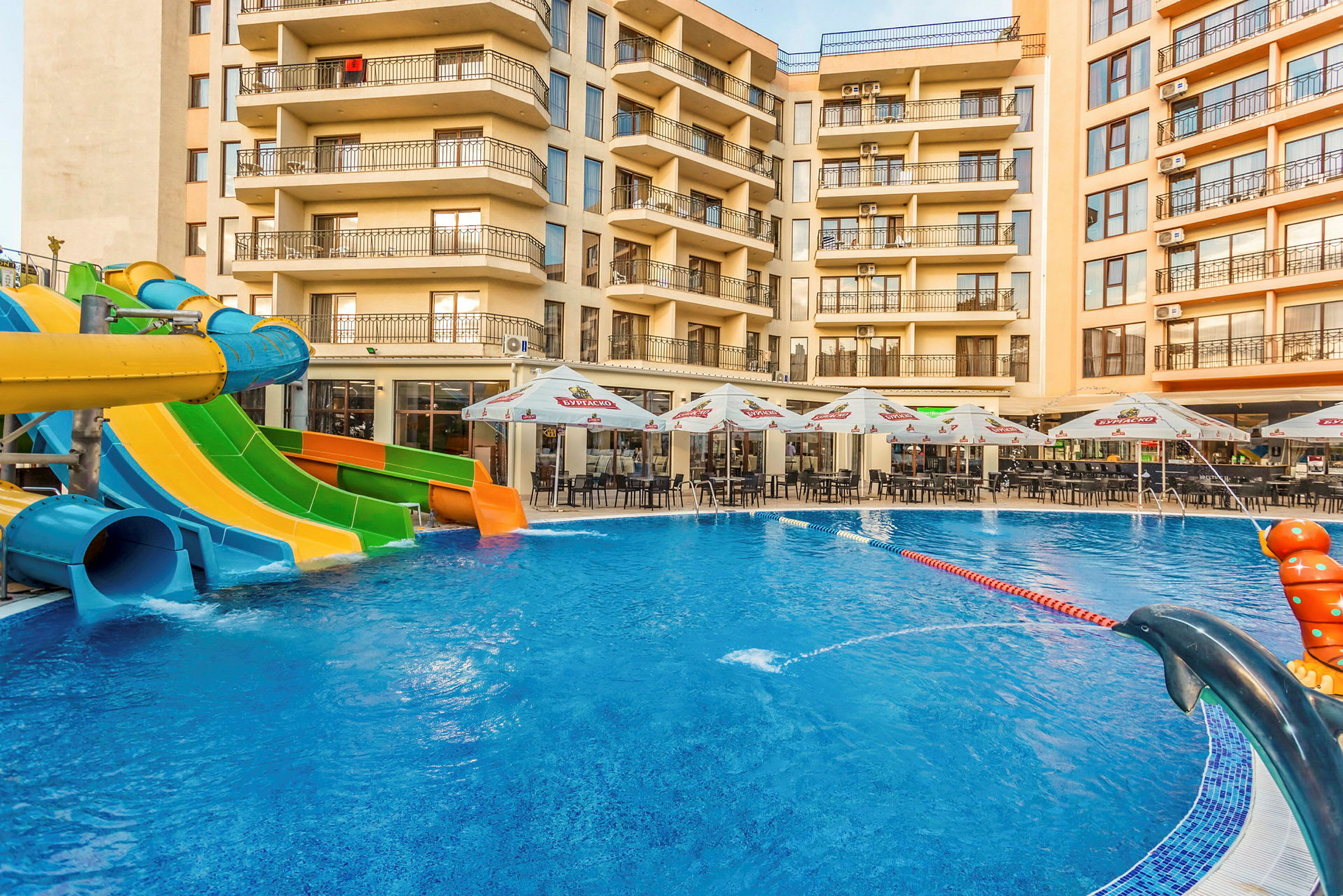 Billedet viser en swimmingpool med vandrutsjebaner i forskellige farver foran et hotel, med udemøbler og parasoller ved poolområdet. (AI-genereret tekst)