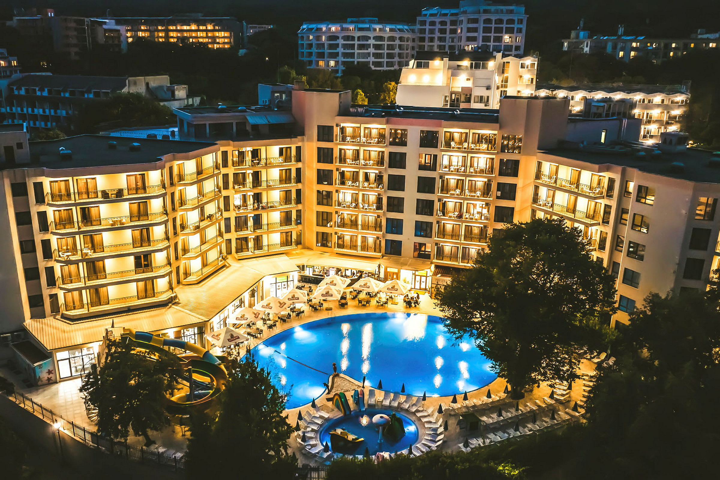 Oplyst hotelbygning med en pool og omkringliggende liggestole, fotograferet om aftenen. (AI-genereret tekst)