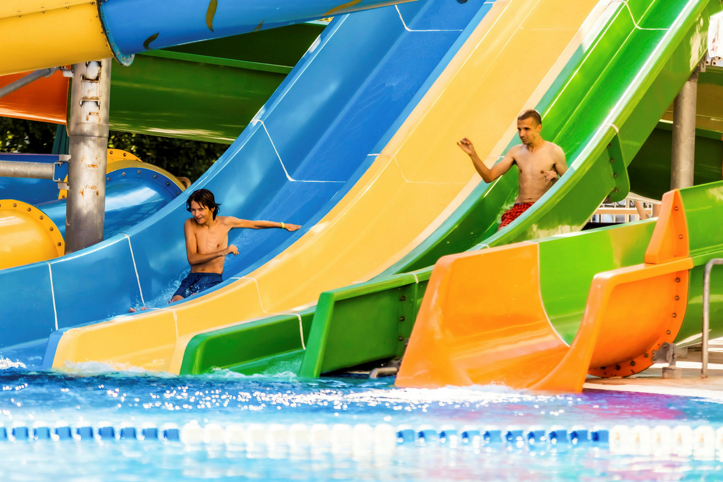 To personer kører ned ad farverige vandrutsjebaner ved en pool. (AI-genereret tekst)