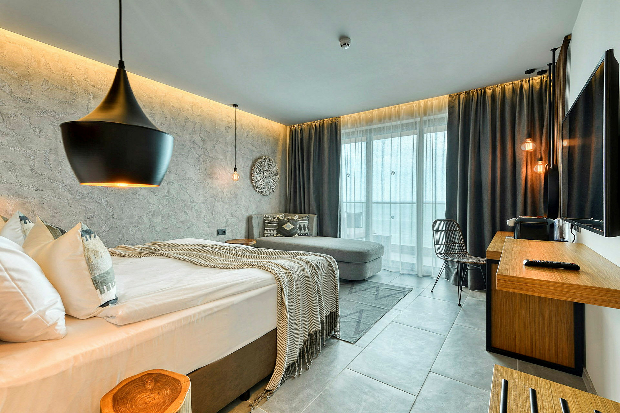 Et moderne hotelværelse med seng, sofa, skrivebord og store vinduer. (AI-genereret tekst)